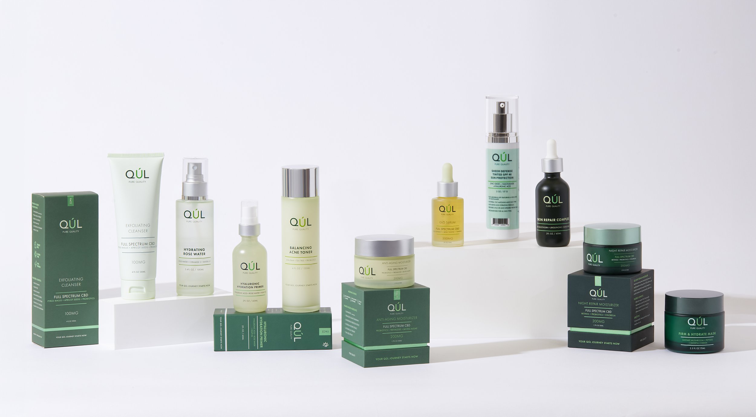 Collection of QÚL skincare products, including exfoliating cleanser, hydrating rose water, balancing acne toner, anti-aging moisturizer, and skin repair serum, arranged on white platforms.