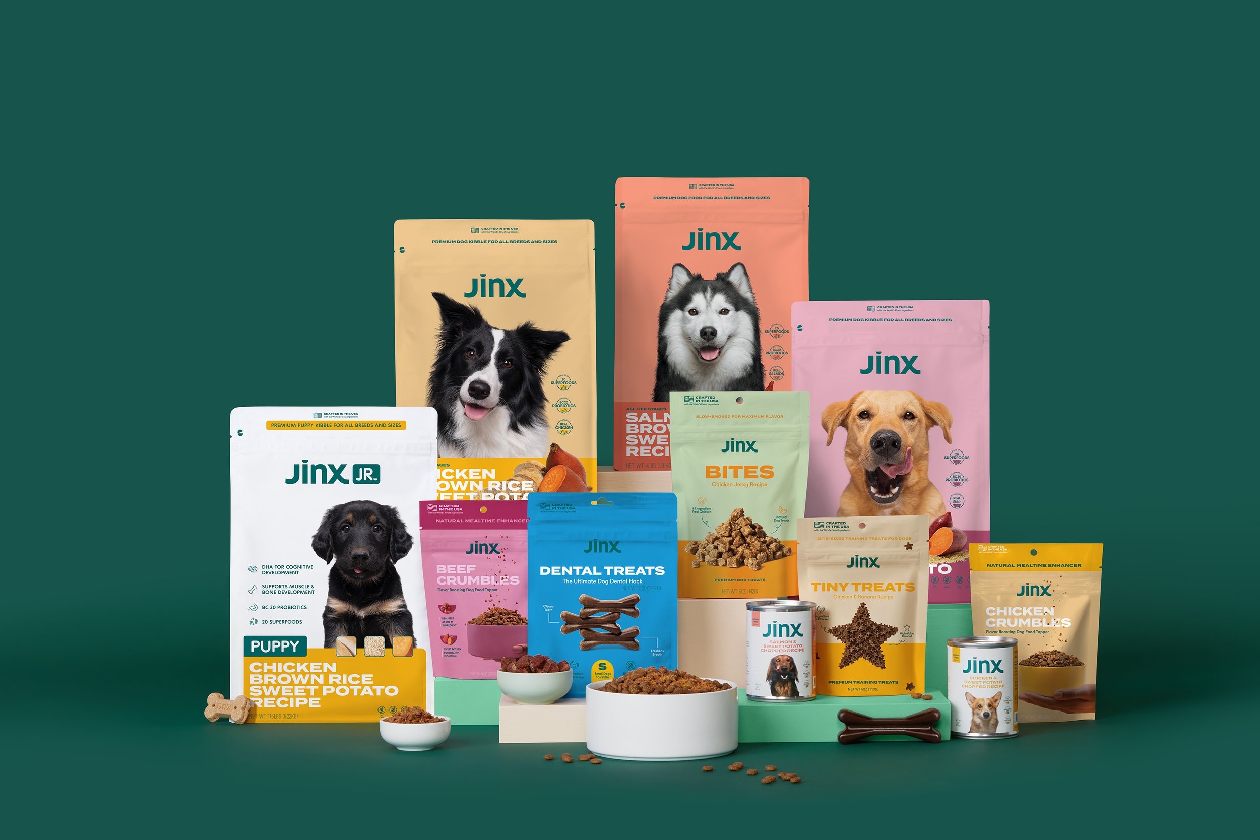 Assorted Jinx dog food and treats on display, including bags of dry food, bites, dental treats, and cans, set against a green background.
