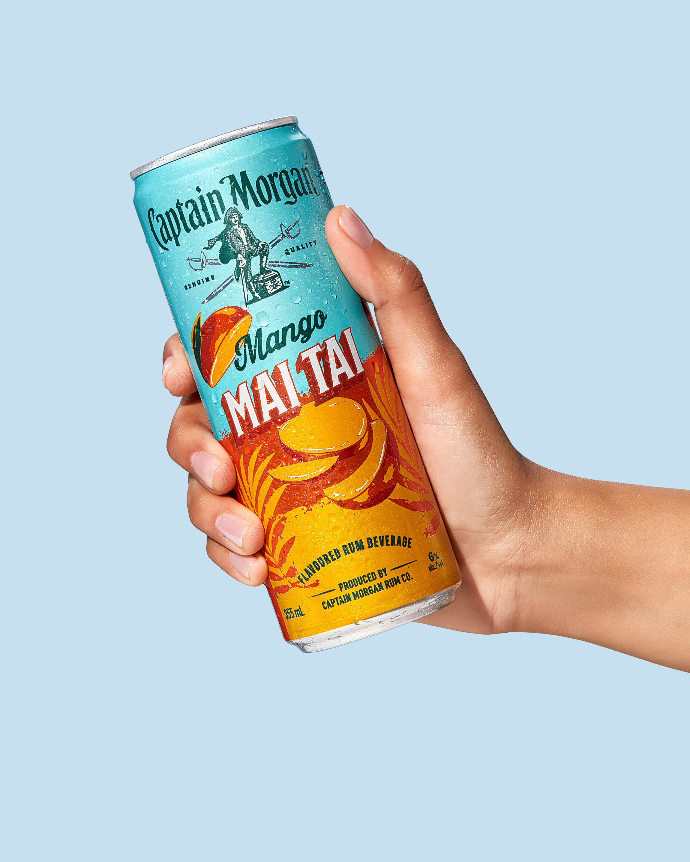 Hand holding Captain Morgan Mango Mai Tai flavored rum beverage can against blue background.