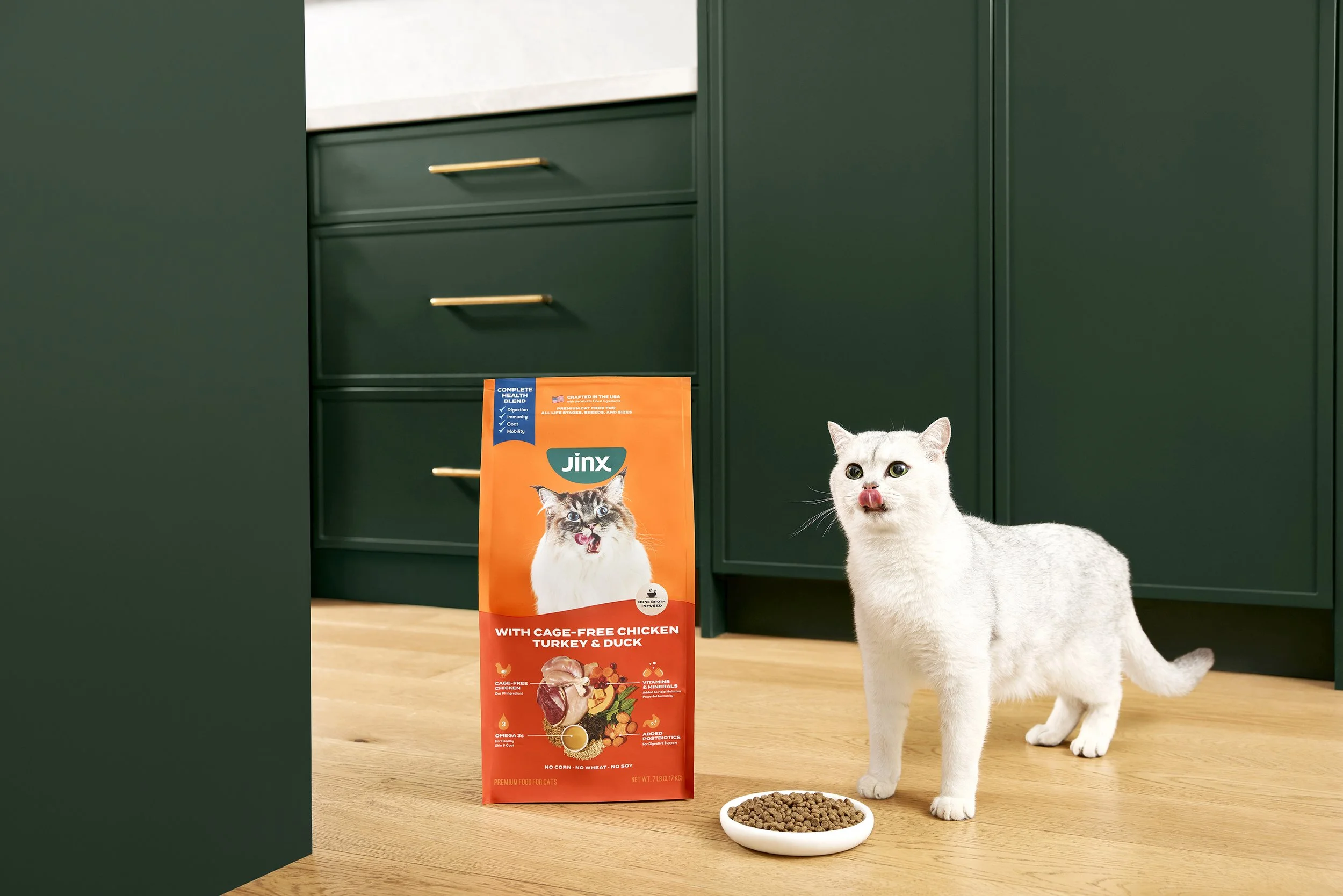 White cat with pink tongue near bowl of dry cat food in kitchen with green cabinets and Jinx cat food bag.