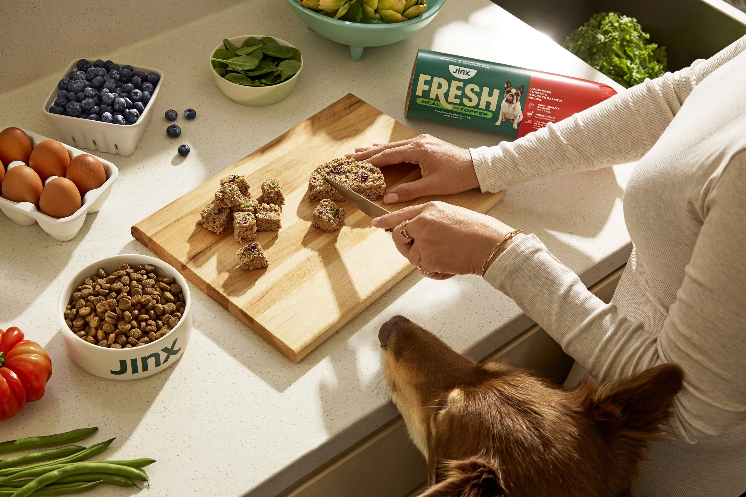 A person chopping homemade dog treats on a wooden cutting board in a kitchen, surrounded by bowls of blueberries, eggs, green beans, and pet food, with a dog looking up at the treats.