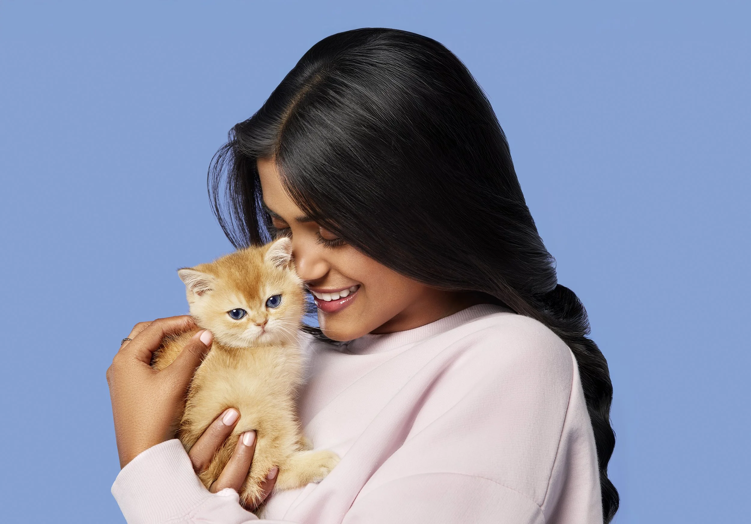 A woman with long black hair smiling and holding an orange kitten with blue eyes against a light blue background.
