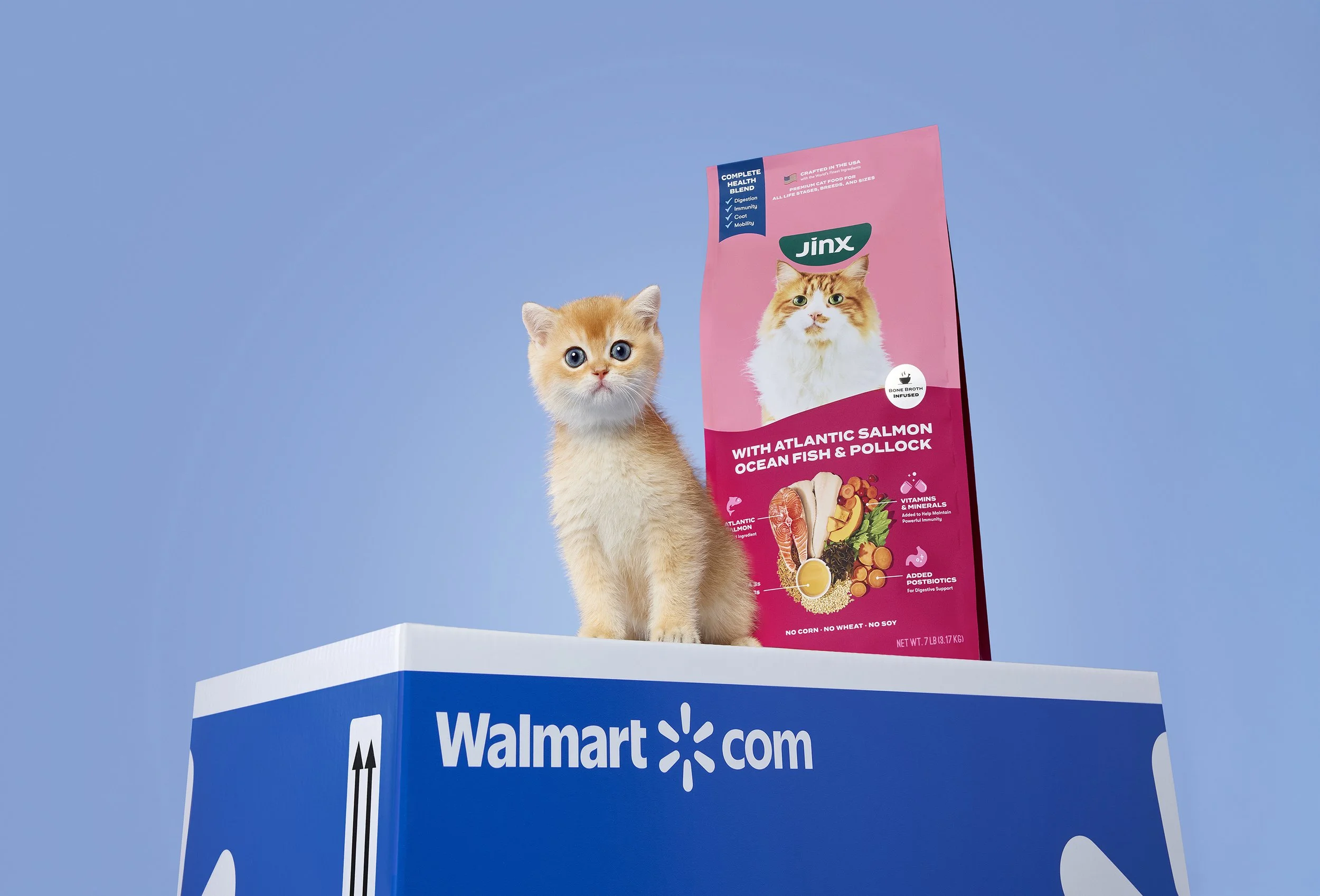 A kitten with orange and white fur sitting on a Walmart box with a bag of Jinx cat food. The cat food bag is pink with an image of a long-haired orange and white cat and features various health benefits.
