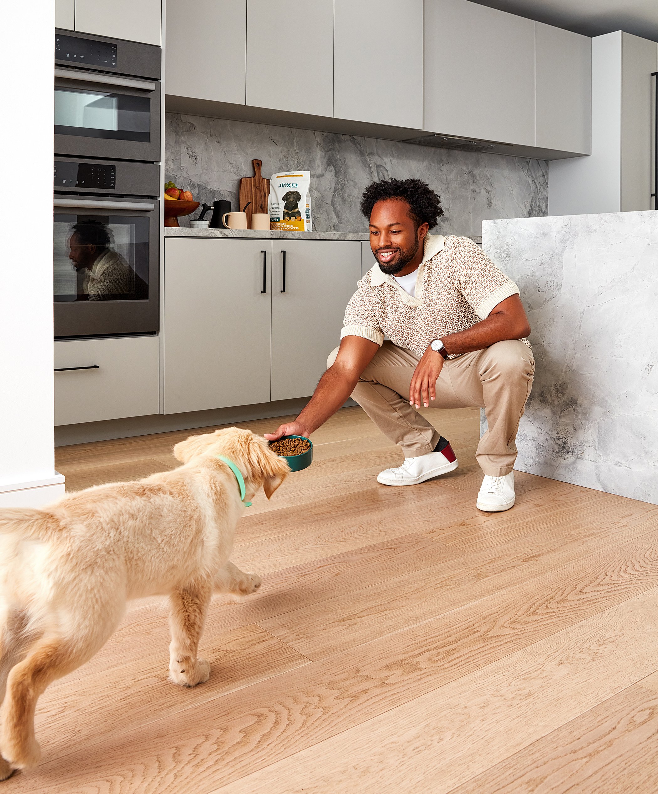 Man feeding a puppy in a modern kitchen with a dog food package on counter.