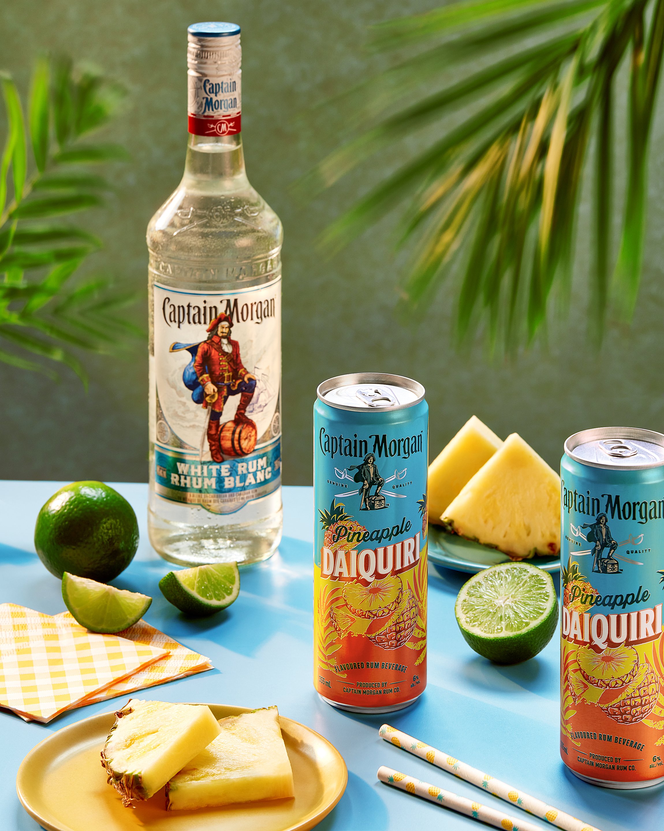 Captain Morgan white rum bottle and two cans of Pineapple Daiquiri, with fresh pineapple slices and lime wedges on a blue surface with tropical plants.