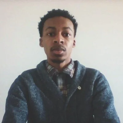 A young man with short curly hair wearing a dark sweater over a plaid shirt, standing indoors against a plain white wall.