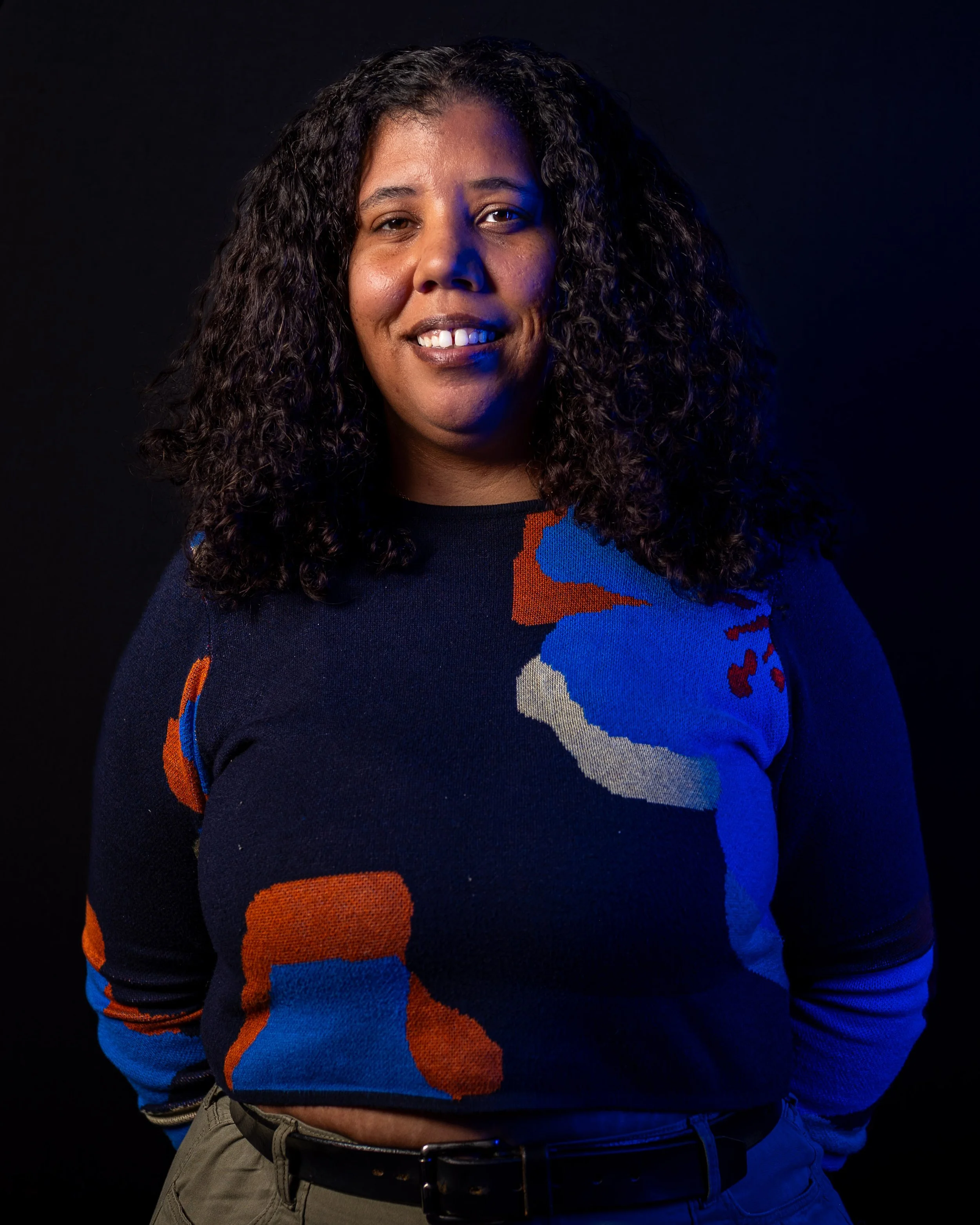 A woman with curly dark hair smiling against a black background. She is wearing a dark sweater with a colorful pattern and gray pants with a black belt.