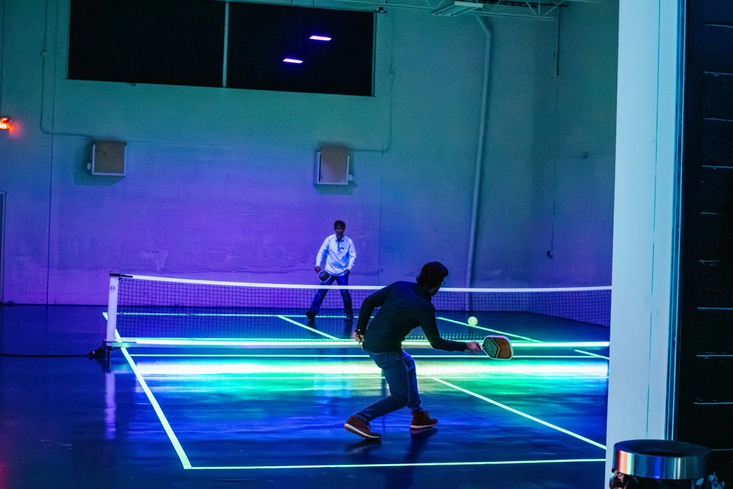 Two people playing pickleball on Golden Hour's indoor court with blue, purple, and green LED lights illuminating the space.