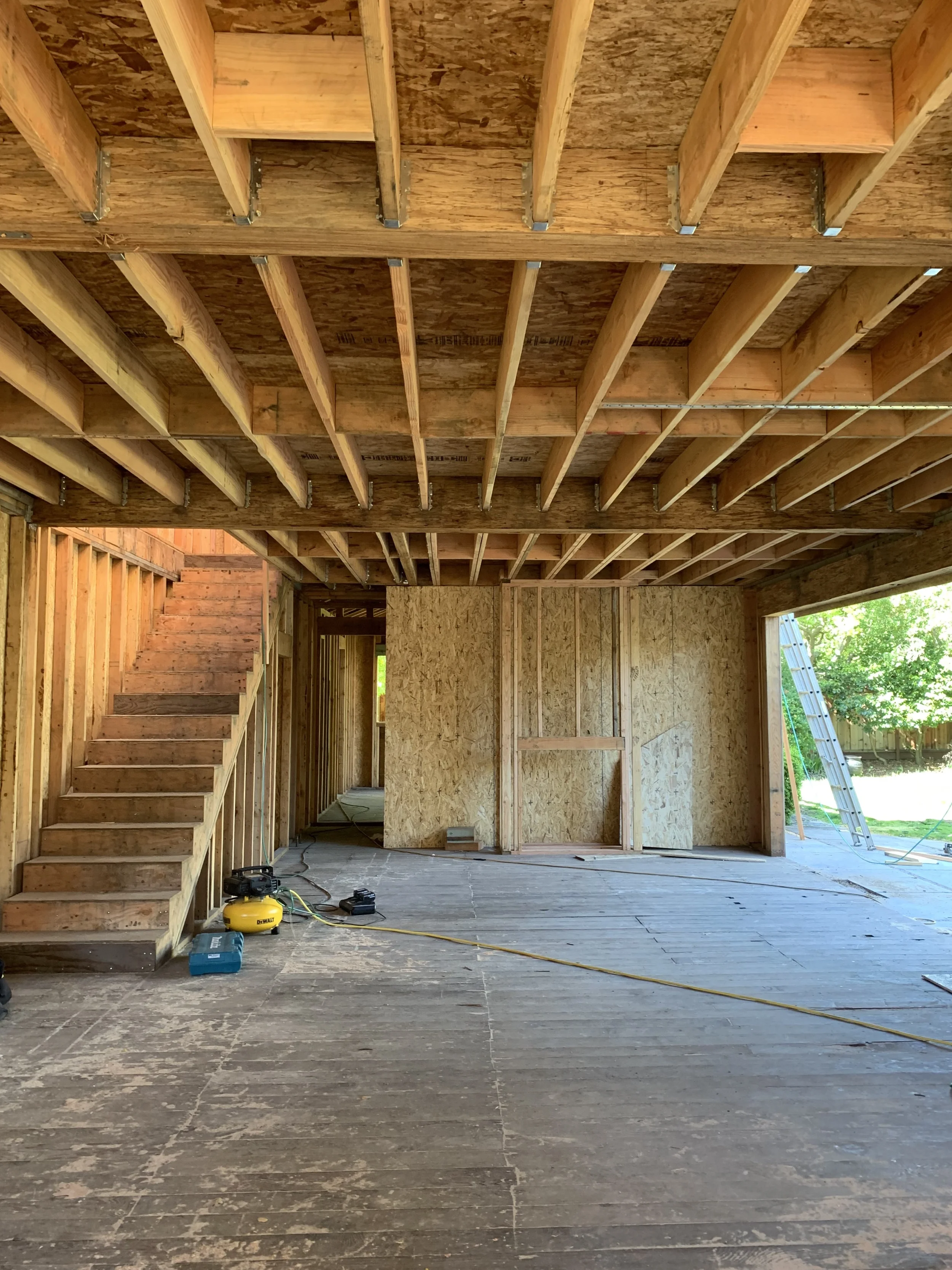 Interior view of a house under construction with exposed wooden framing, stairs on the left, and a ladder outside through an opening on the right.