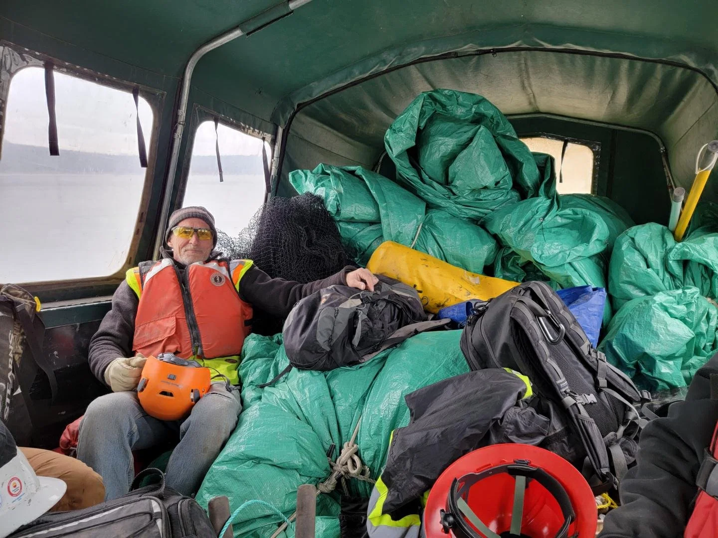 A man wearing a red safety vest, yellow safety glasses, and a beanie hat sitting in a truck filled with green tarp-covered equipment, backpacks, and safety gear, with water and a boat in the background.