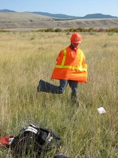 A person in an orange safety vest and cap walking through a grassy field, with a backpack on the ground nearby and open land with hills in the background.