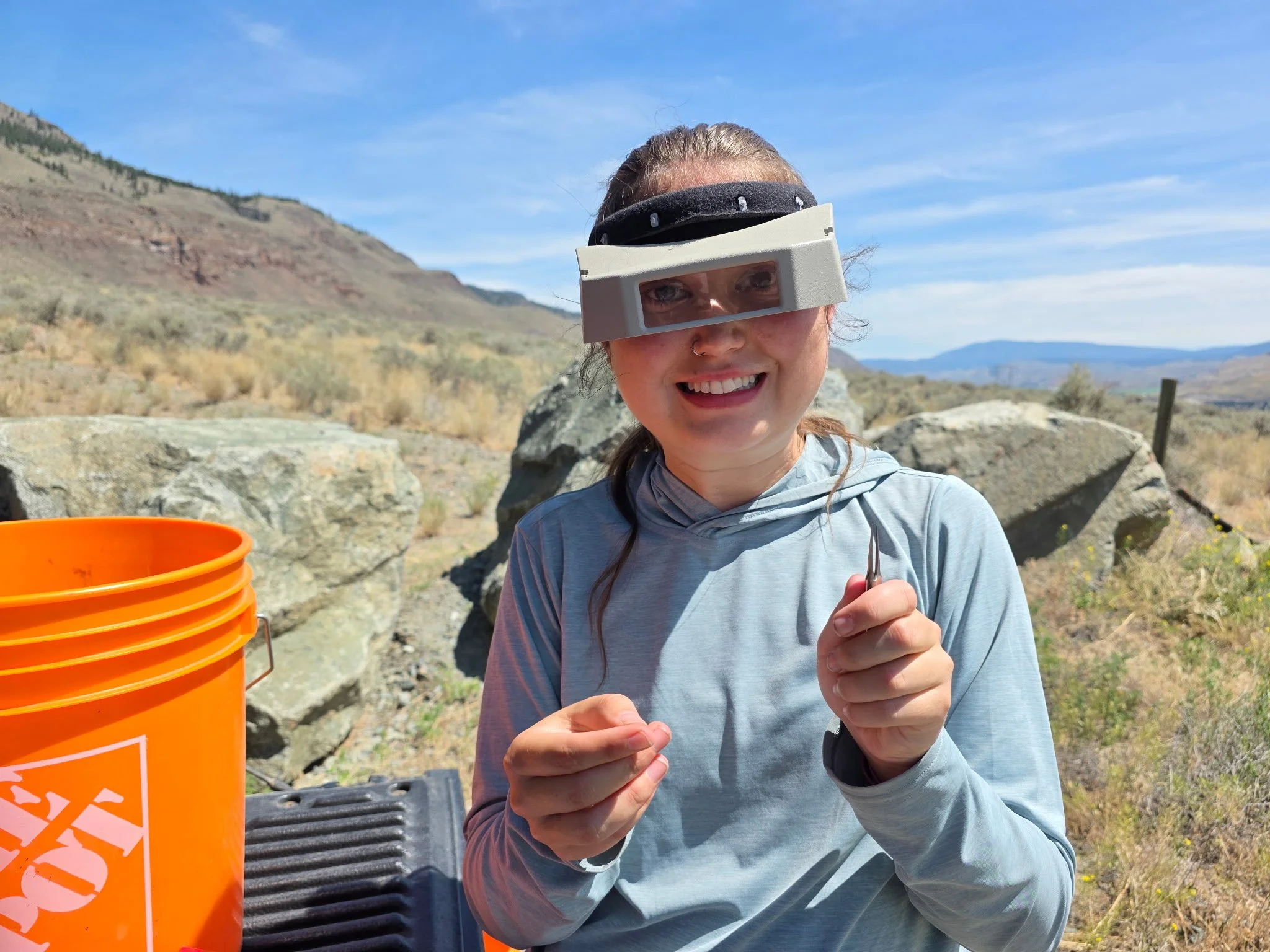 A woman outdoors in a mountainous area wearing protective glasses, smiling, and holding a small object in her hands. An orange bucket is visible nearby.