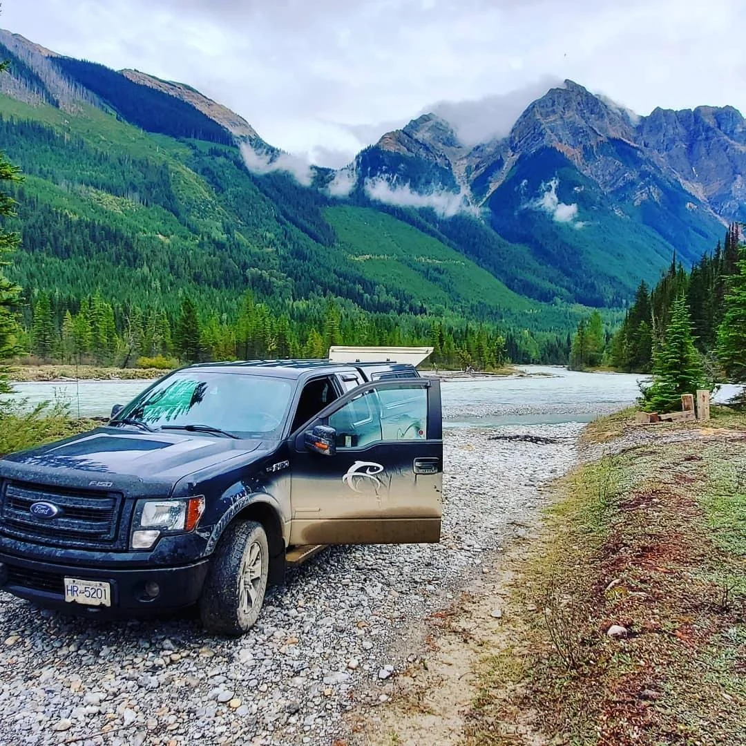 A black Ford truck parked on a gravel area near a river with green forest and tall mountains in the background.