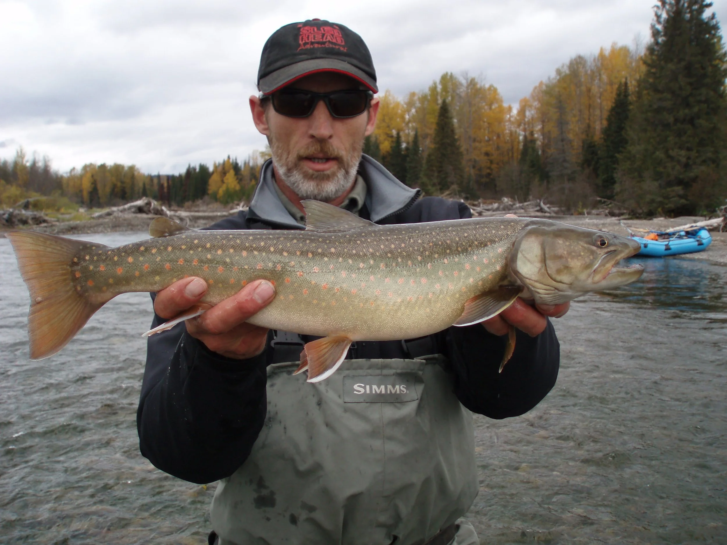 A man wearing sunglasses and outdoor gear holding a large fish with a patterned body. The background shows a river with an autumnal forest and kayaks on the riverbank.