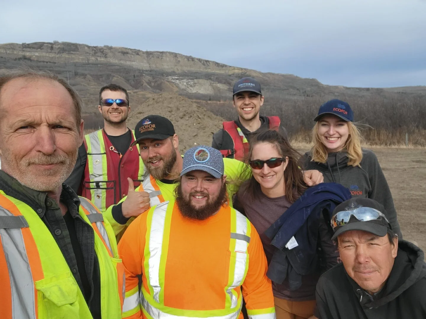 Group of nine people outdoors in a rural area with hills in the background, smiling and wearing safety gear including vests, hats, and sunglasses.
