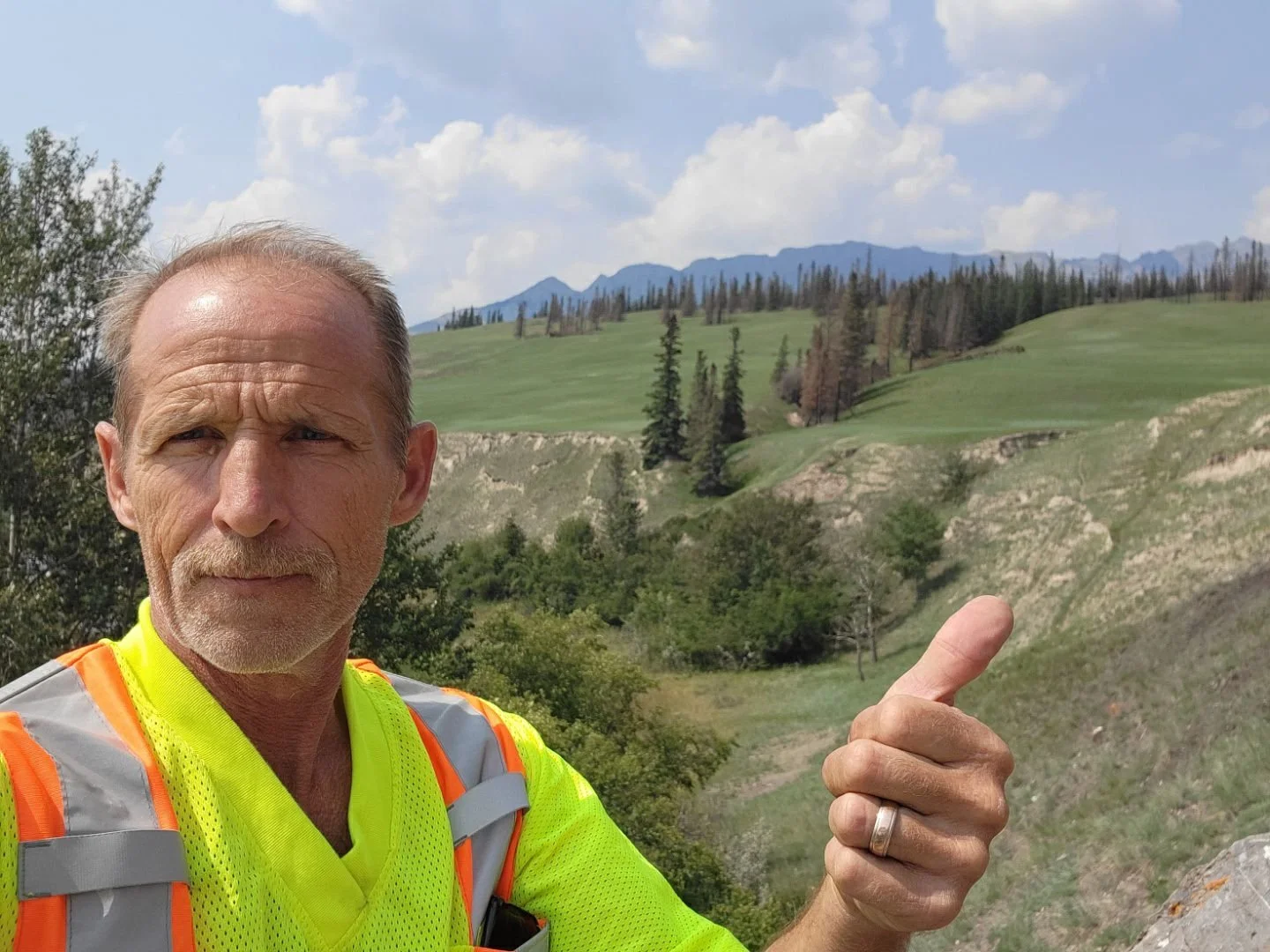 A man in a neon yellow shirt and safety vest giving a thumbs up in front of a scenic mountainous landscape with grassy hills, trees, and mountains in the background.