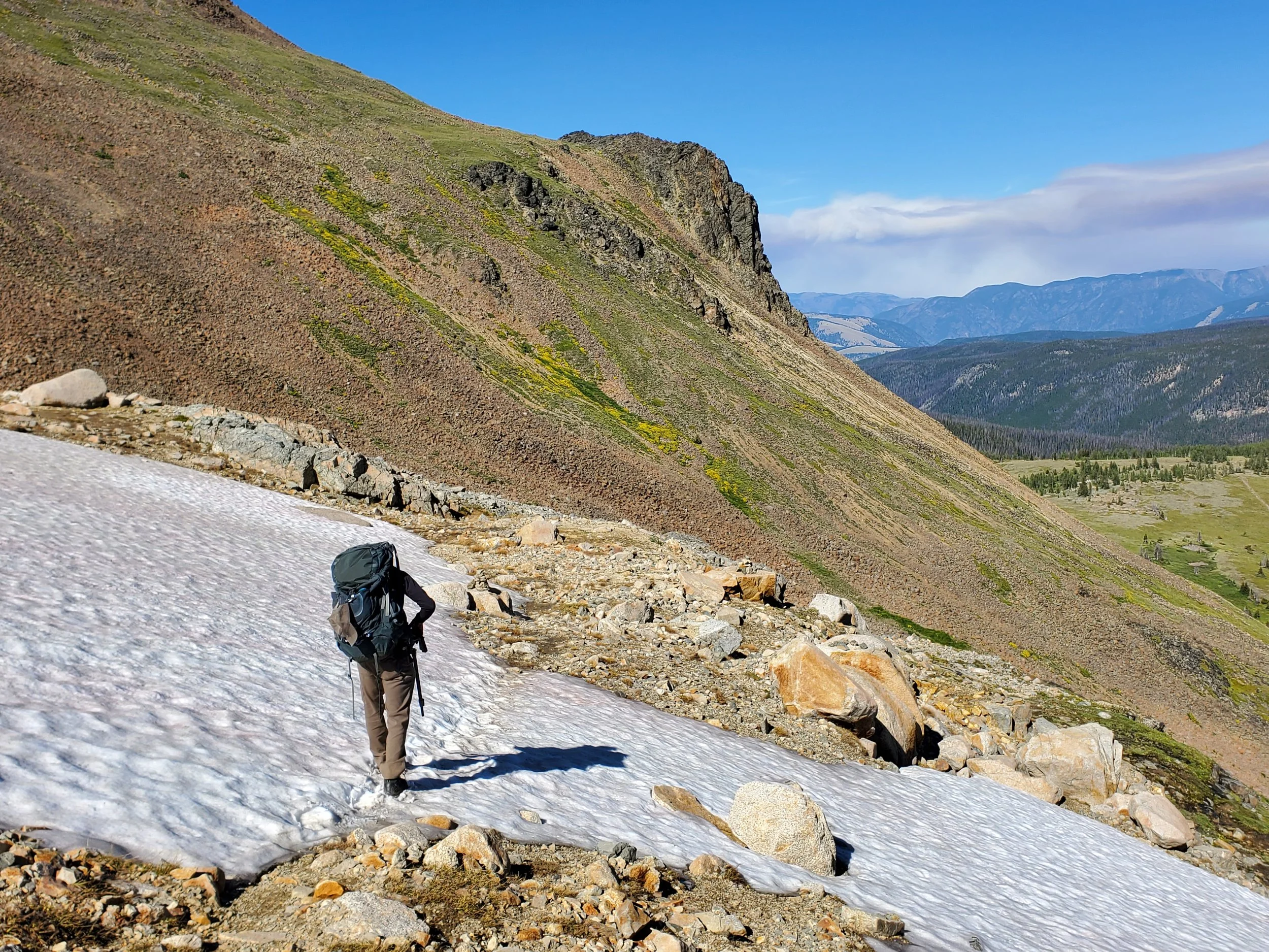 A hiker with a backpack walking on a snow-covered trail in a mountainous landscape with rocky slopes and distant mountain ranges under a partly cloudy sky.