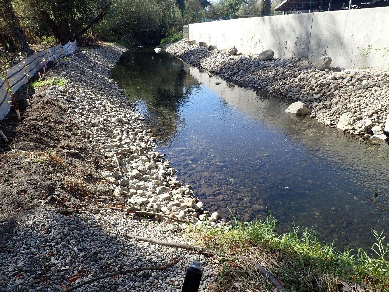 A small, shallow creek with clear water flowing through a rocky and gravel-lined channel, bordered by a concrete wall on one side and trees on the other.