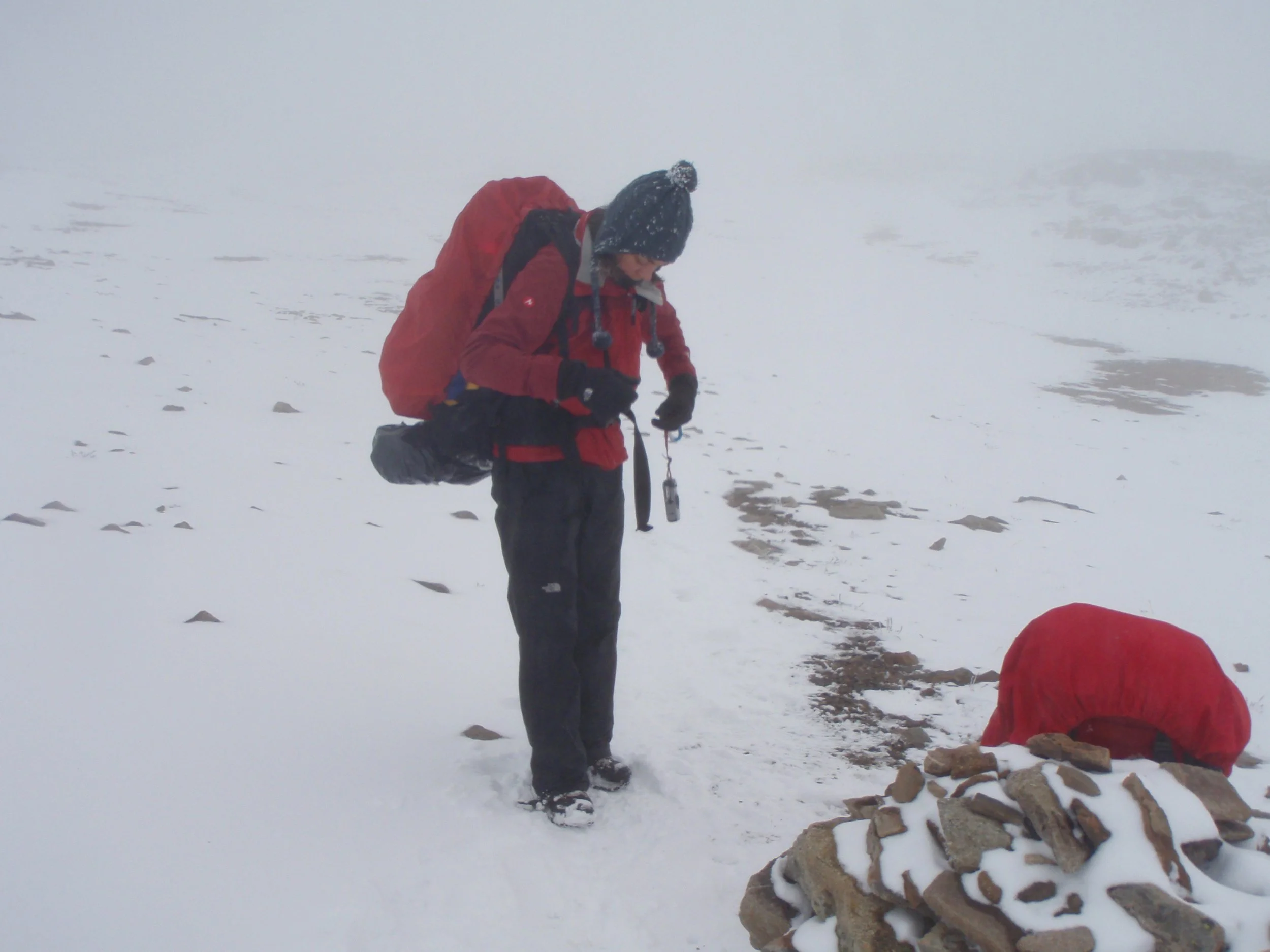 Person dressed in red and black outdoor clothing, wearing a black hat with a pom-pom, adjusting equipment in a snowy, foggy landscape. Another person in red is kneeling on the snow near a pile of rocks, possibly setting up a cairn or marker.
