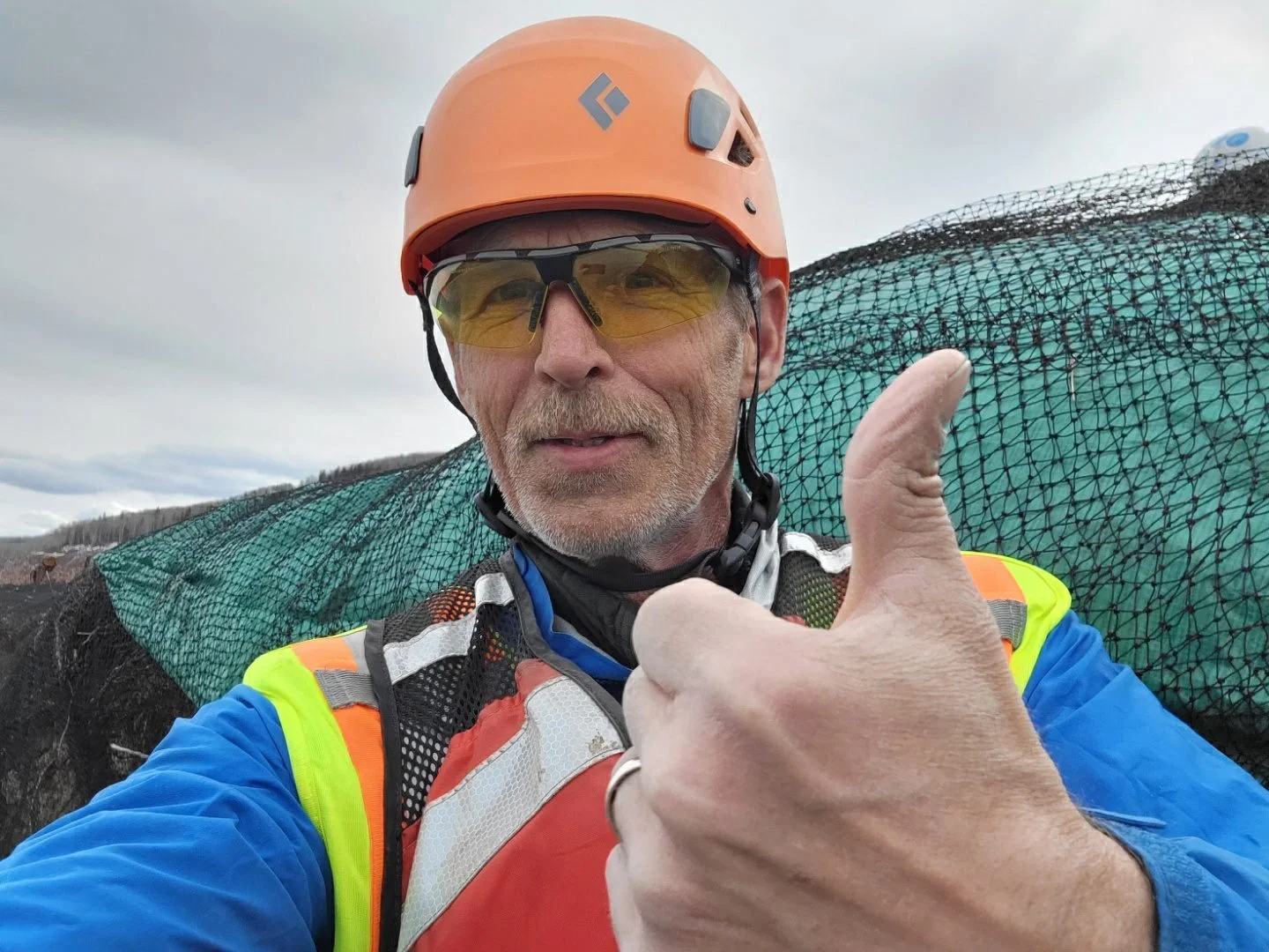 A man wearing an orange safety helmet, yellow-tinted safety glasses, and a high-visibility jacket taking a selfie with a thumbs-up gesture, with a green netted barrier in the background outdoors.