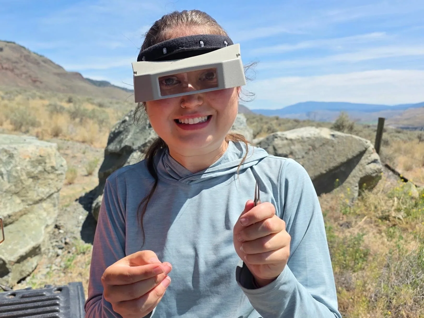 A woman outdoors with mountains and rocks in the background, wearing sunglasses and a headband, smiling, holding a small tool or object.