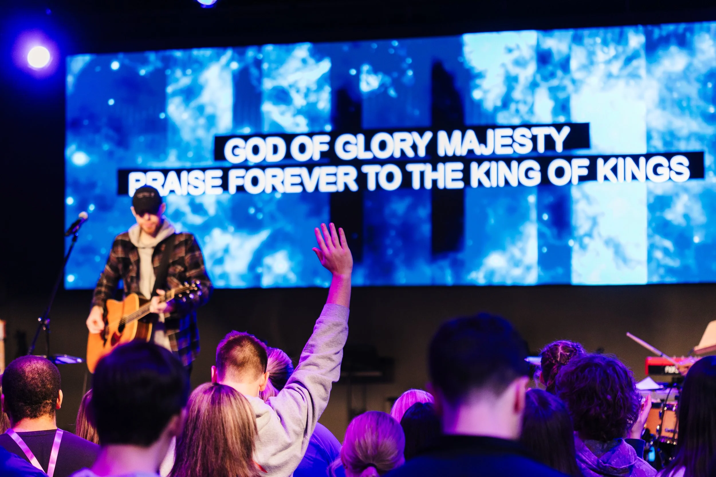 A live church or concert event with a man playing a guitar on stage. A large screen in the background displays the words 'GOD OF GLORY MAJESTY PRAISE FOREVER TO THE KING OF KINGS'. Audience members have raised hands in the foreground.