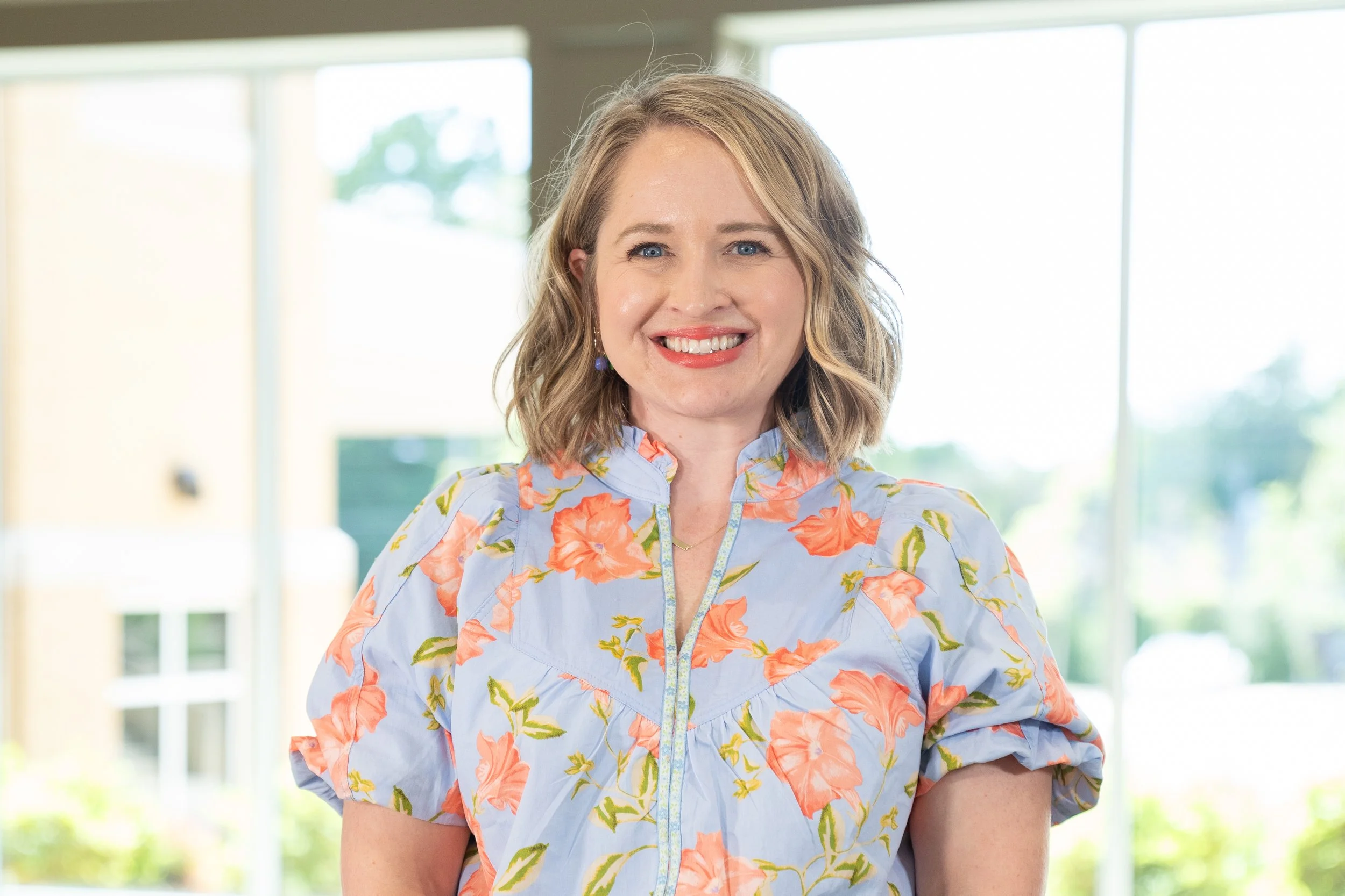 A smiling woman with shoulder-length blonde hair, wearing a light blue floral blouse, standing in front of large windows with a view of trees outside.