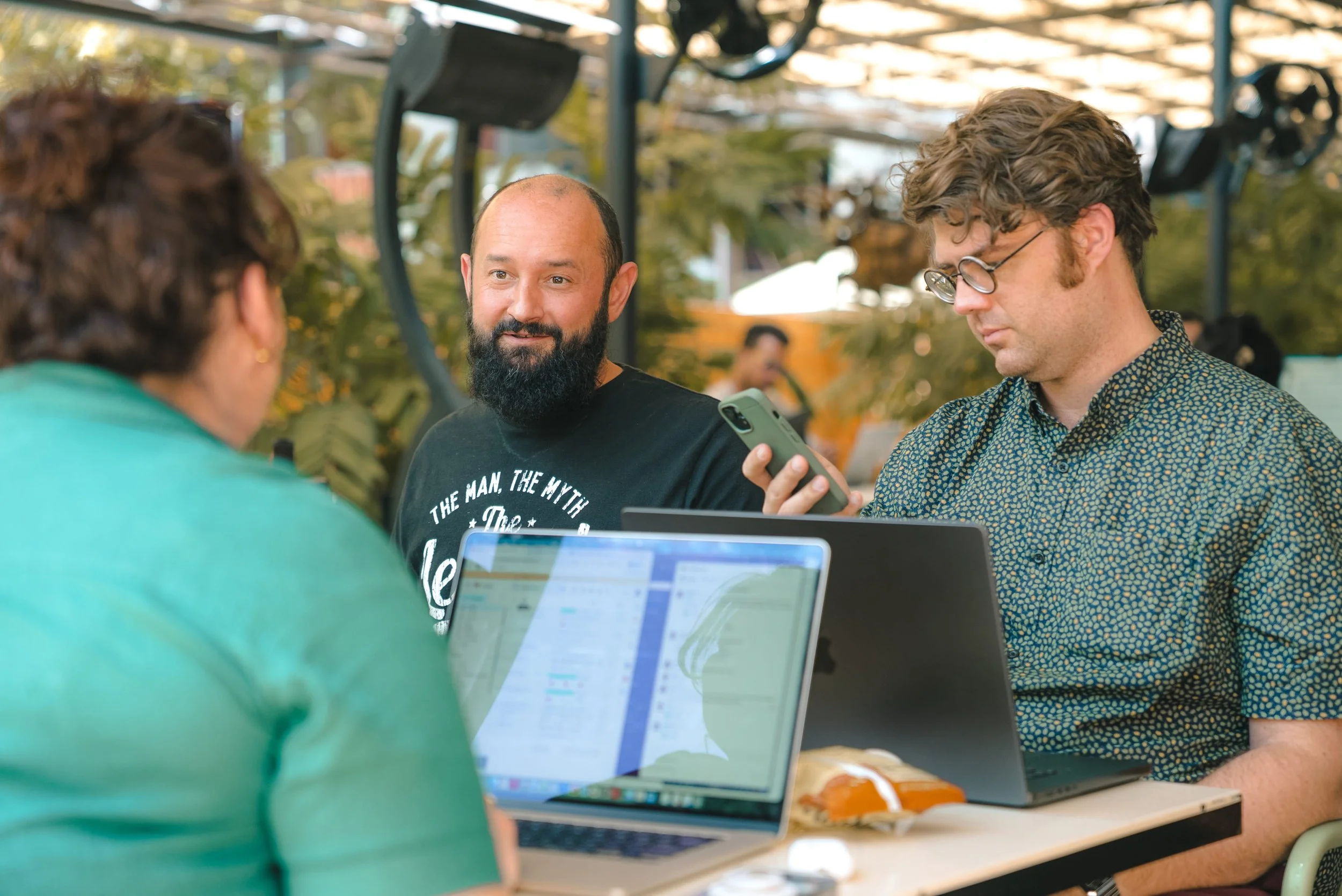 A group of young adults gathered around a laptop, engaging in a discussion or collaboration outdoors with trees and a canopy overhead.