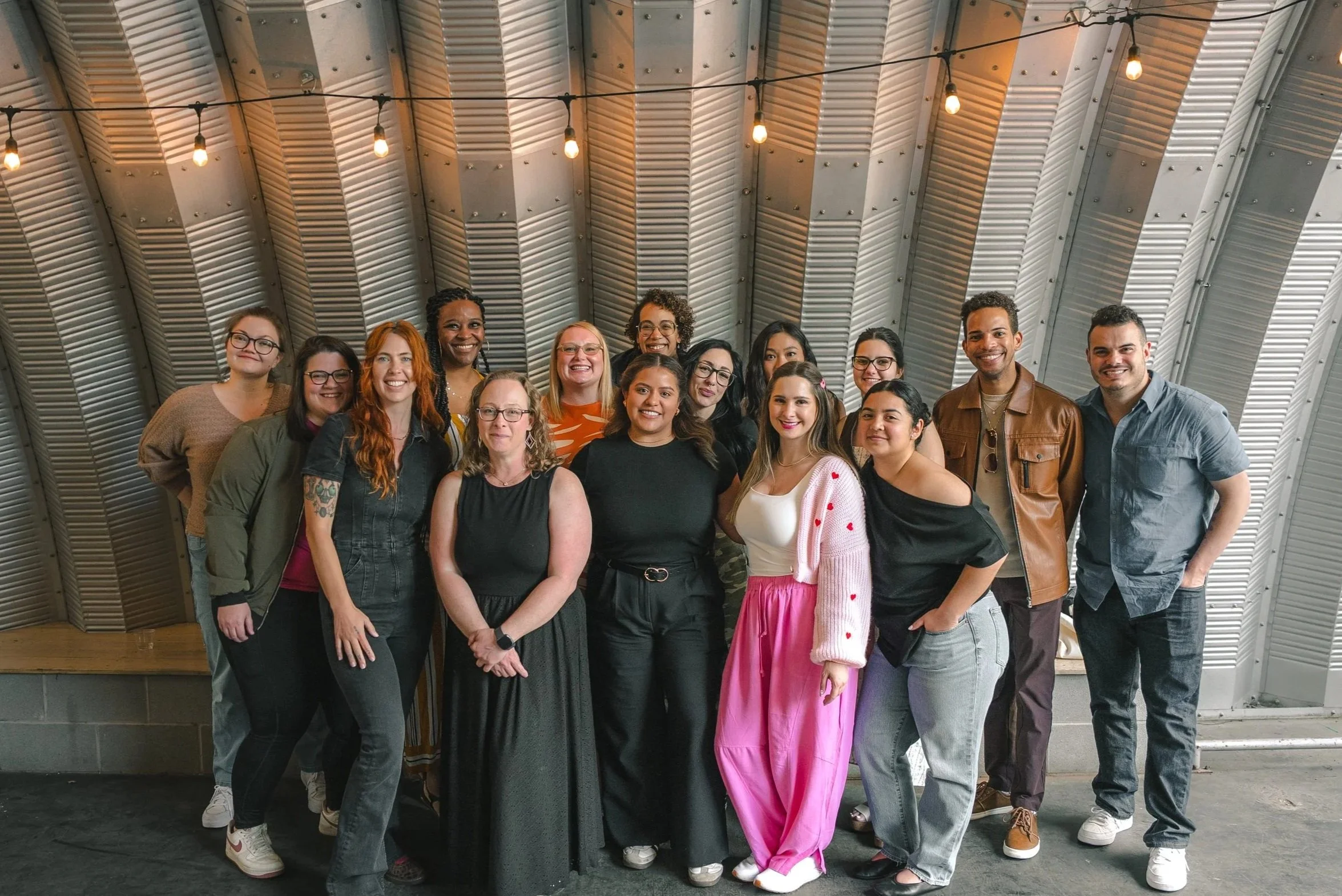 A diverse group of people standing together indoors under a vaulted ceiling with hanging lights, smiling at the camera.