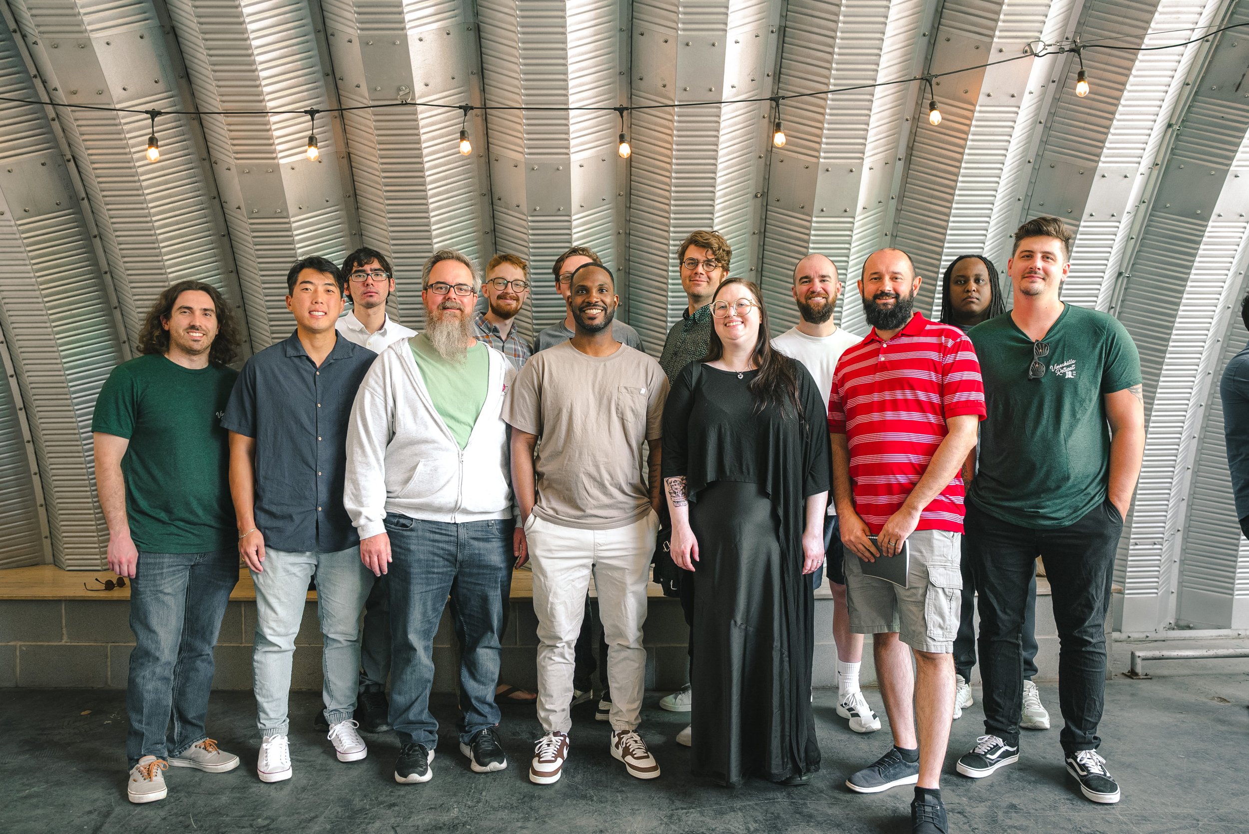 Group of diverse people standing together indoors under a curved metal roof with string lights.