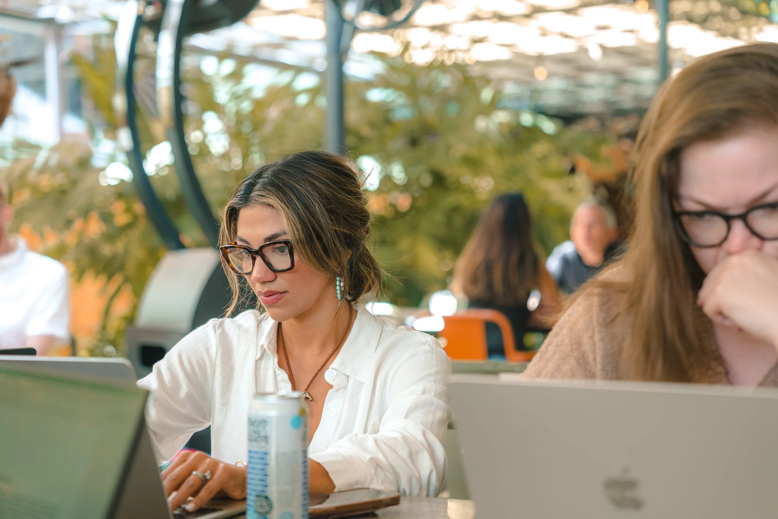 A woman with glasses and earrings working on a laptop at a desk in an open, glass-enclosed space with people and greenery in the background.