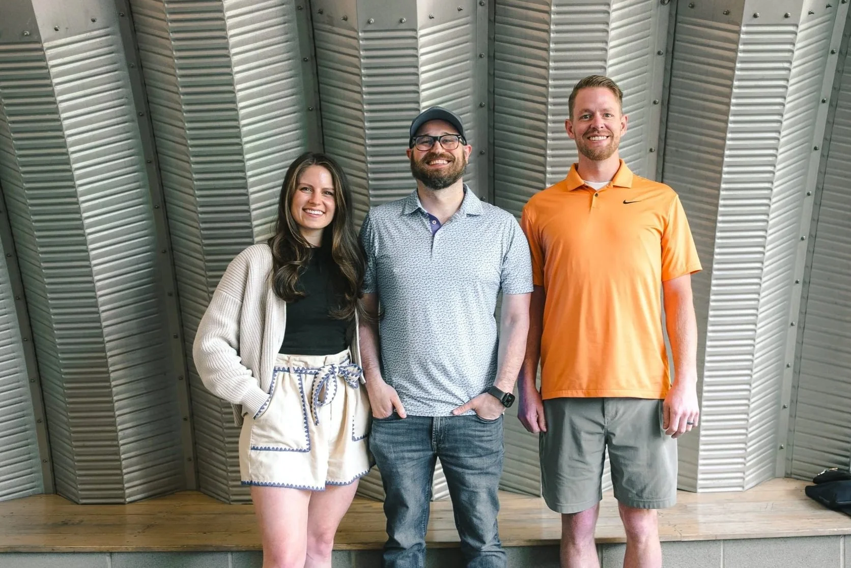 Three smiling people standing together indoors in front of a metallic wall. The woman on the left has long brown hair and is wearing a black top, cream-colored shorts with blue trim, and a cream cardigan. The man in the middle has a beard, glasses, a cap, and is wearing a patterned short-sleeved shirt and jeans. The man on the right has short hair, a beard, and is wearing an orange Nike polo shirt and khaki shorts.