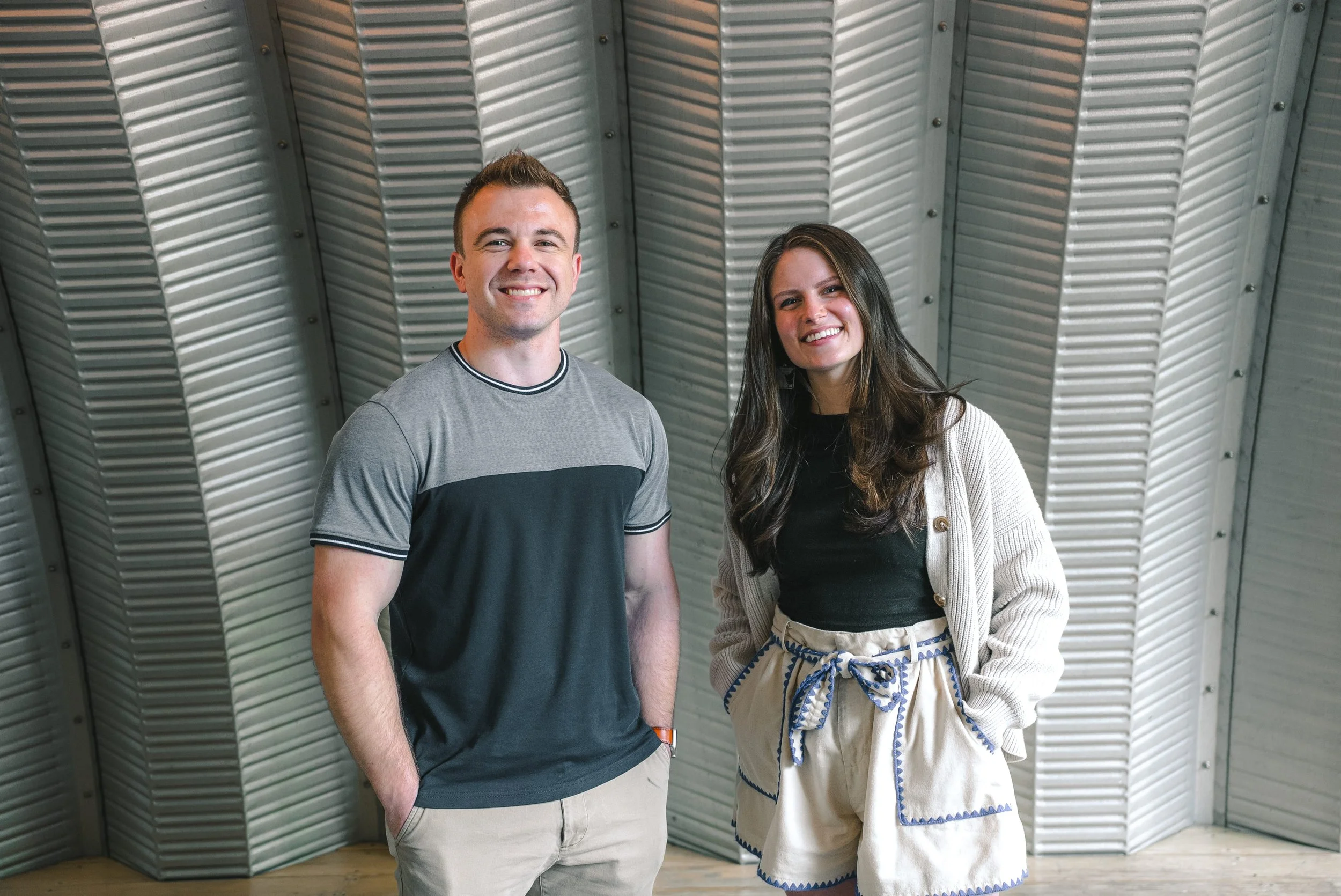 A smiling young man and woman standing in front of a metallic wall, dressed casually.