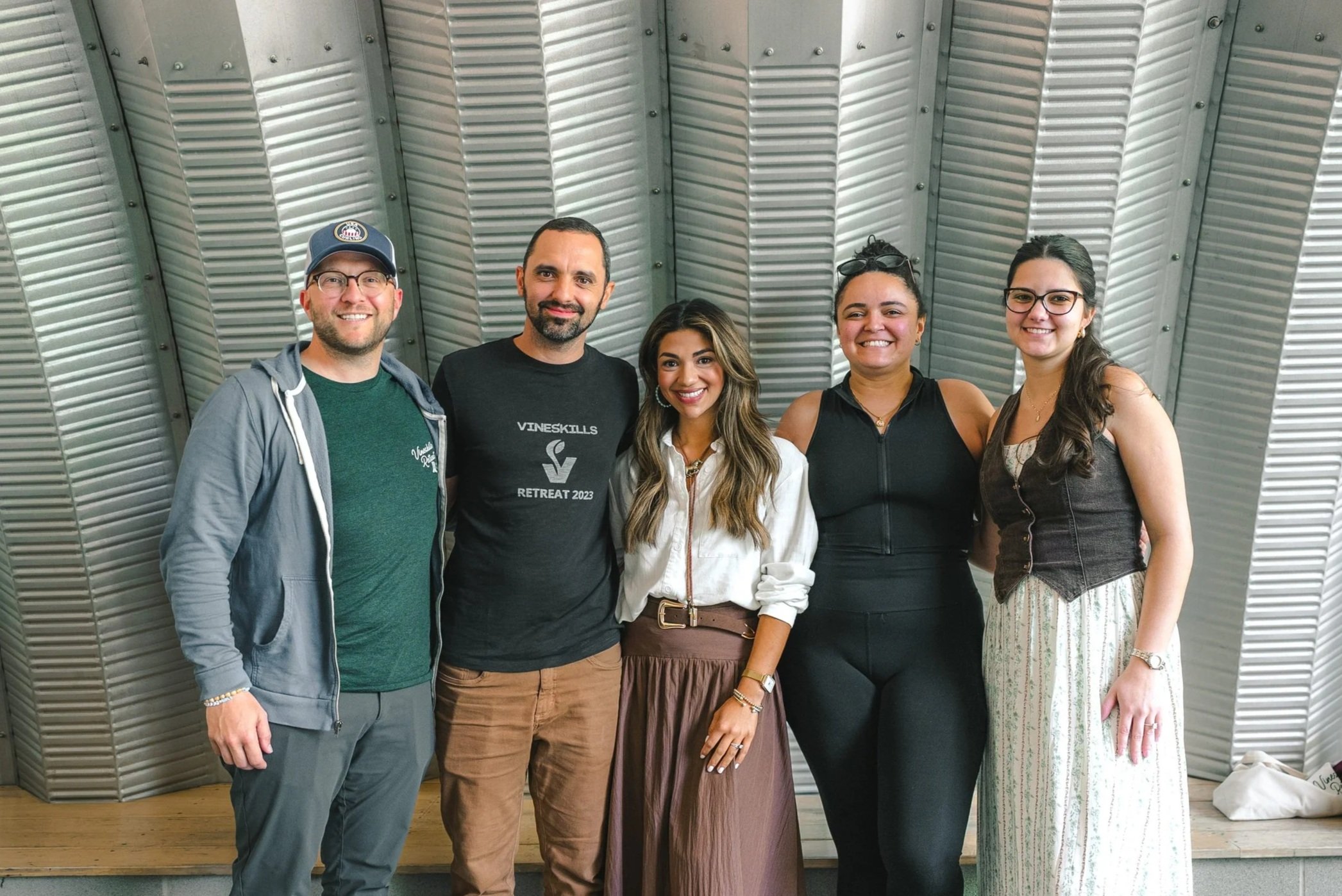 Group of five diverse people standing close together, smiling, in front of a metallic, curved wall at an indoor event.