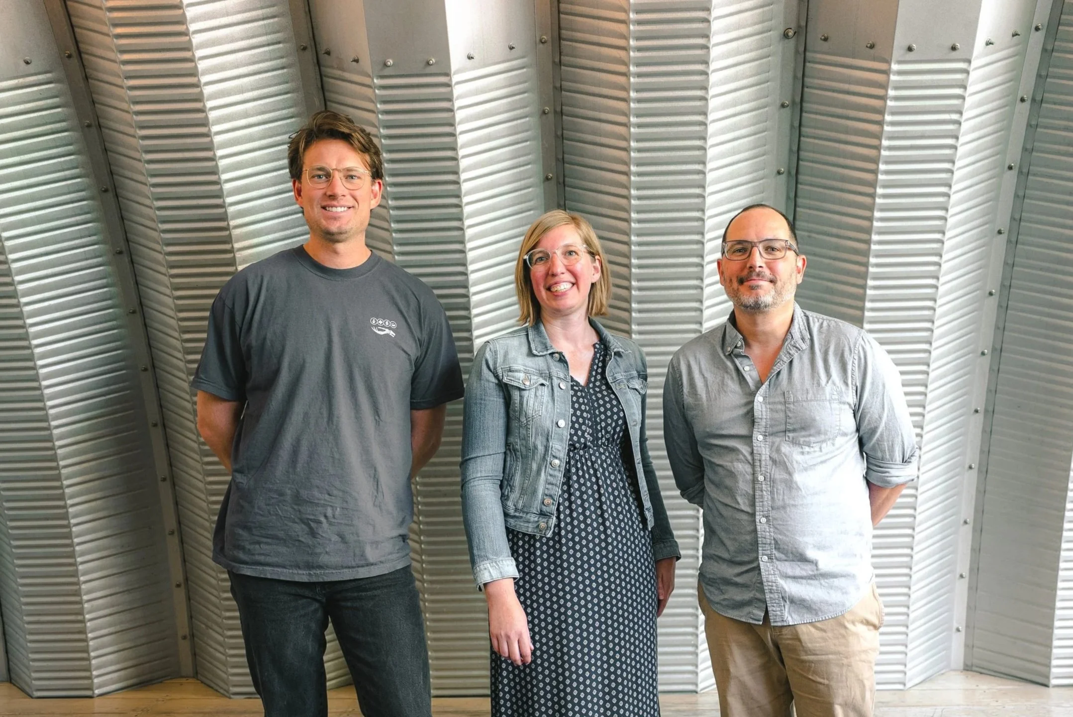 Three people standing side by side in front of a metallic, ribbed wall. The person on the left is a man with glasses and short dark hair, wearing a gray t-shirt. The person in the middle is a woman with shoulder-length blonde hair, wearing glasses, a denim jacket, and a dark polka dot dress. The person on the right is a man with glasses and short hair, wearing a gray button-up shirt and khakis.
