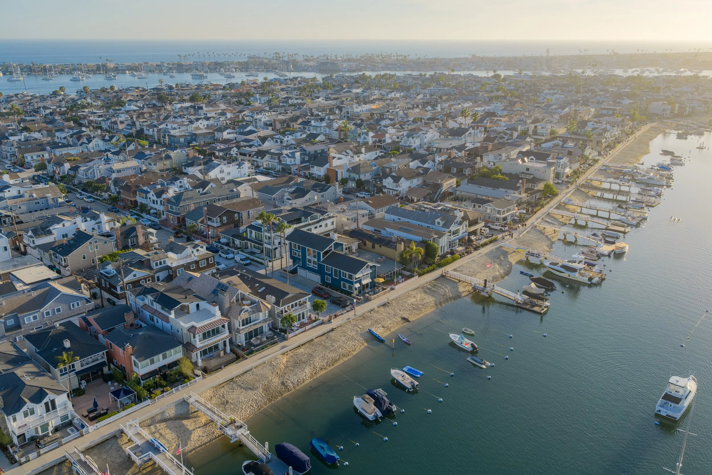 An aerial view of a coastal residential area with houses along a marina, boats docked in the water, and a distant ocean with sailboats and palm trees.