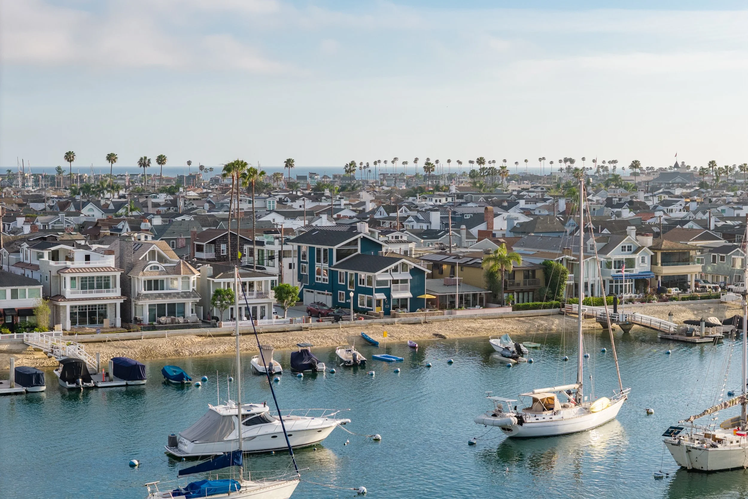 A coastal neighborhood with houses lining the waterfront and sailboats docked in a marina with palm trees in the background.
