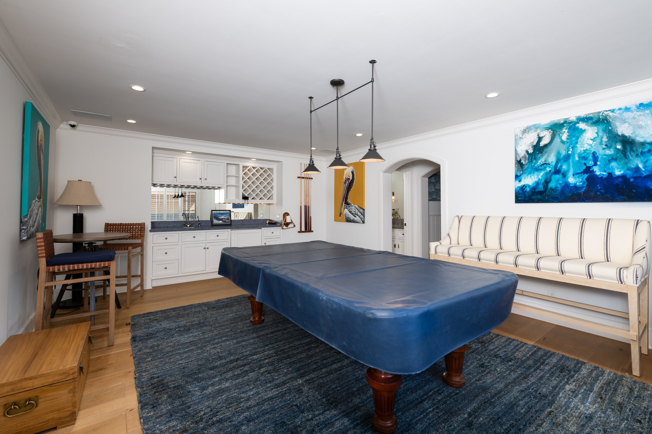 Living room with a large abstract blue painting on the wall, a striped sofa, a dining table with a blue cover, and wall art of a pelican. There are kitchen cabinets and a black countertop in the background.
