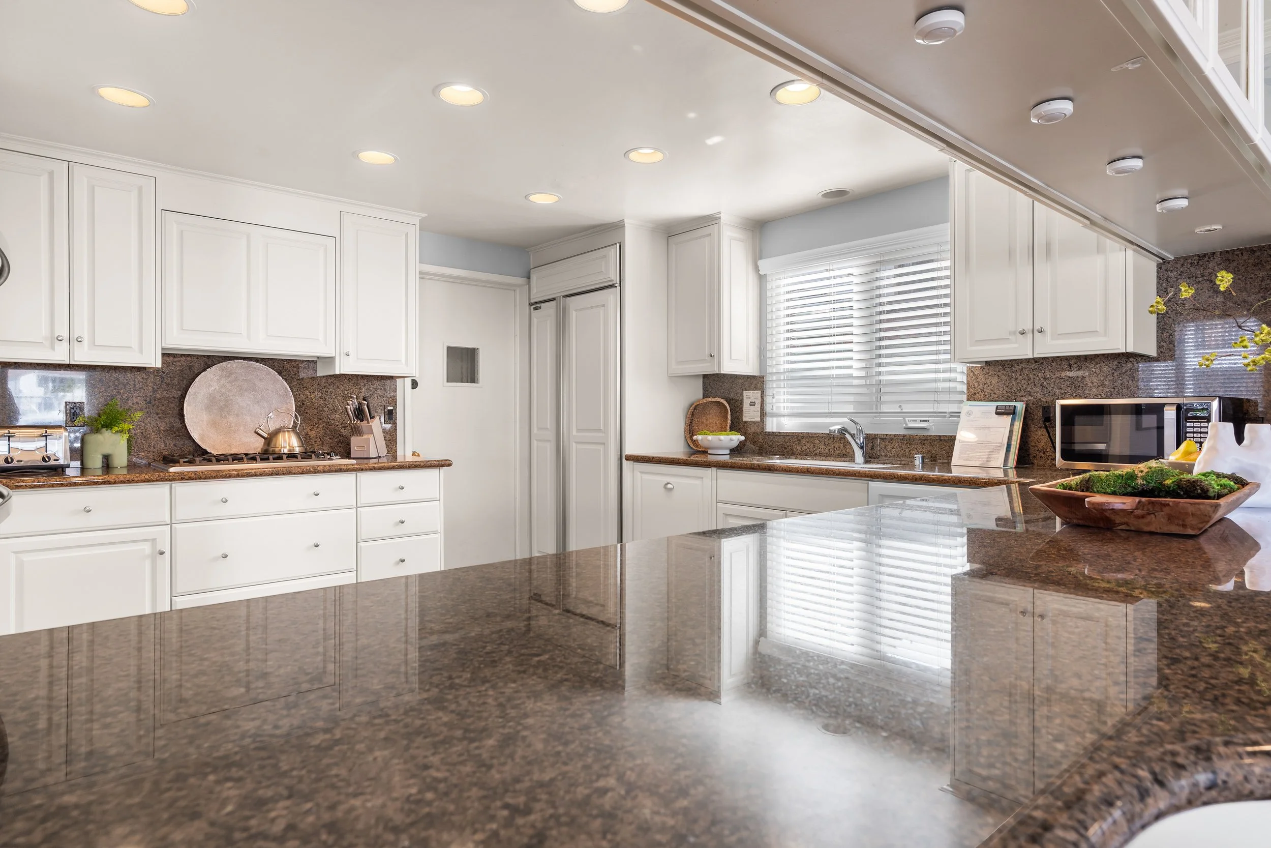 A modern kitchen with white cabinets, brown granite countertops, a window with white blinds, and various kitchen appliances and decor.