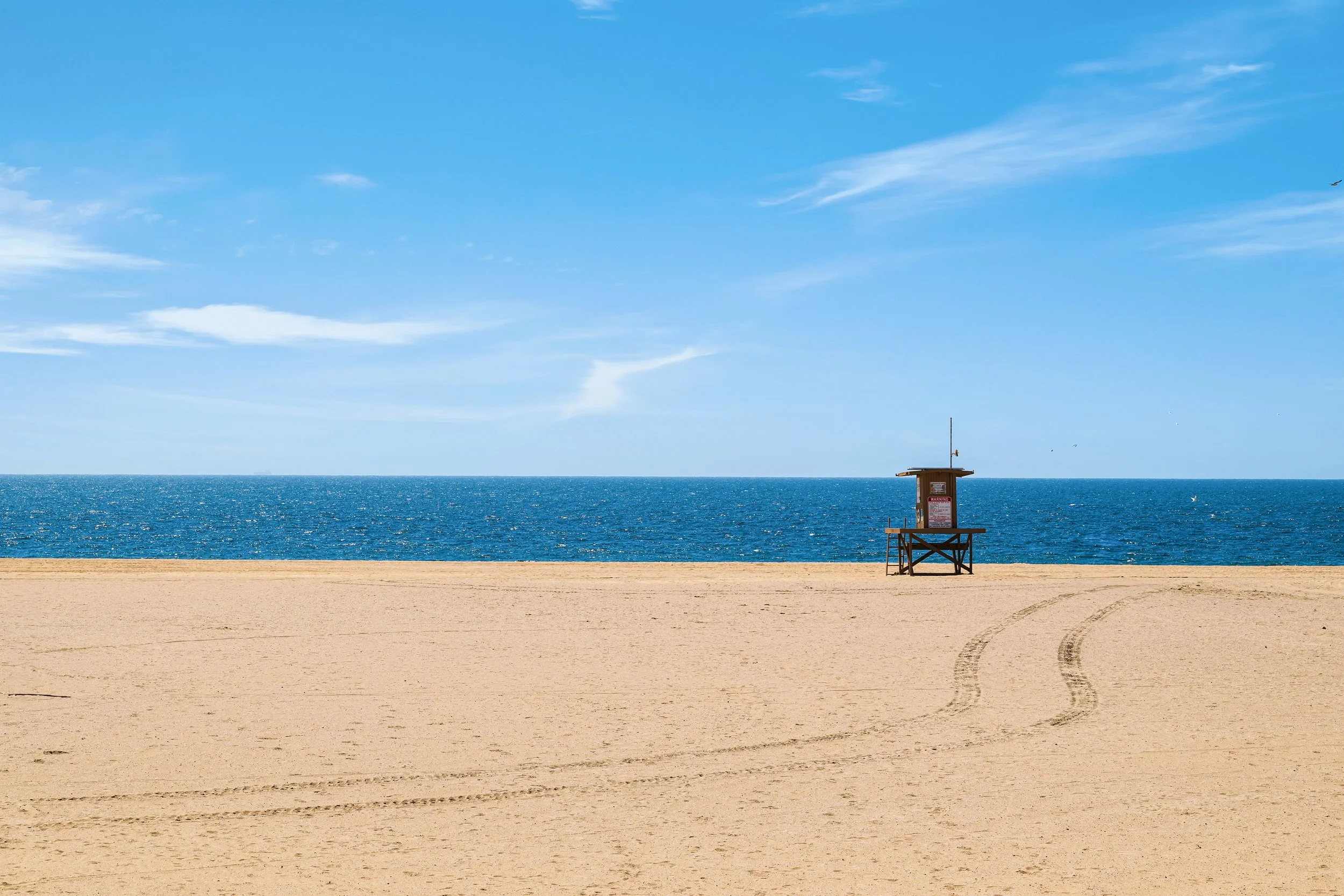 A sandy beach with a lifeguard tower, calm blue ocean water, and a bright blue sky with some clouds.