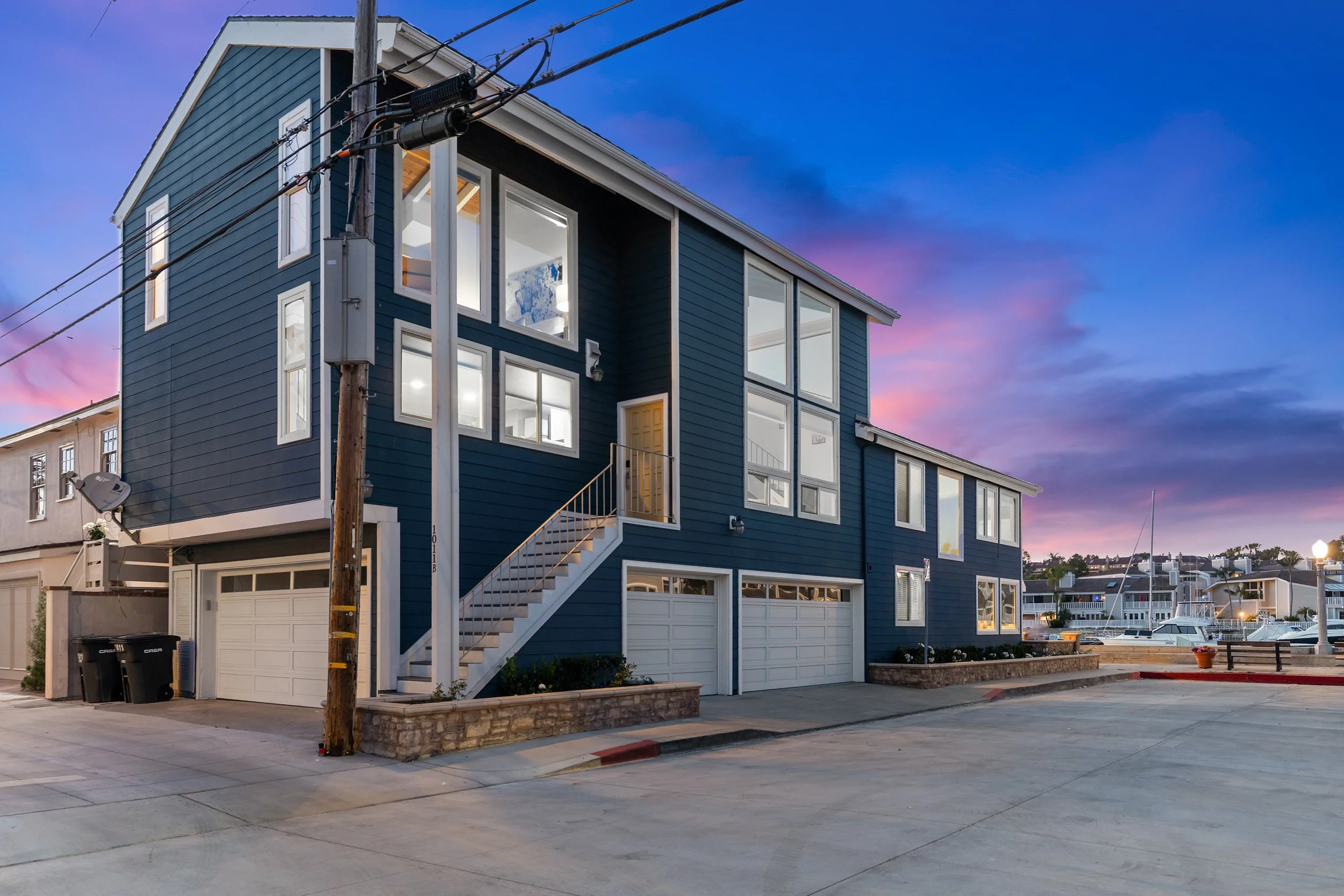 A blue multi-story house with large windows on each floor, a staircase leading to the front door, and a garage on the ground level. The house is near a body of water with boats and other houses visible in the background, under a sunset sky with purpl