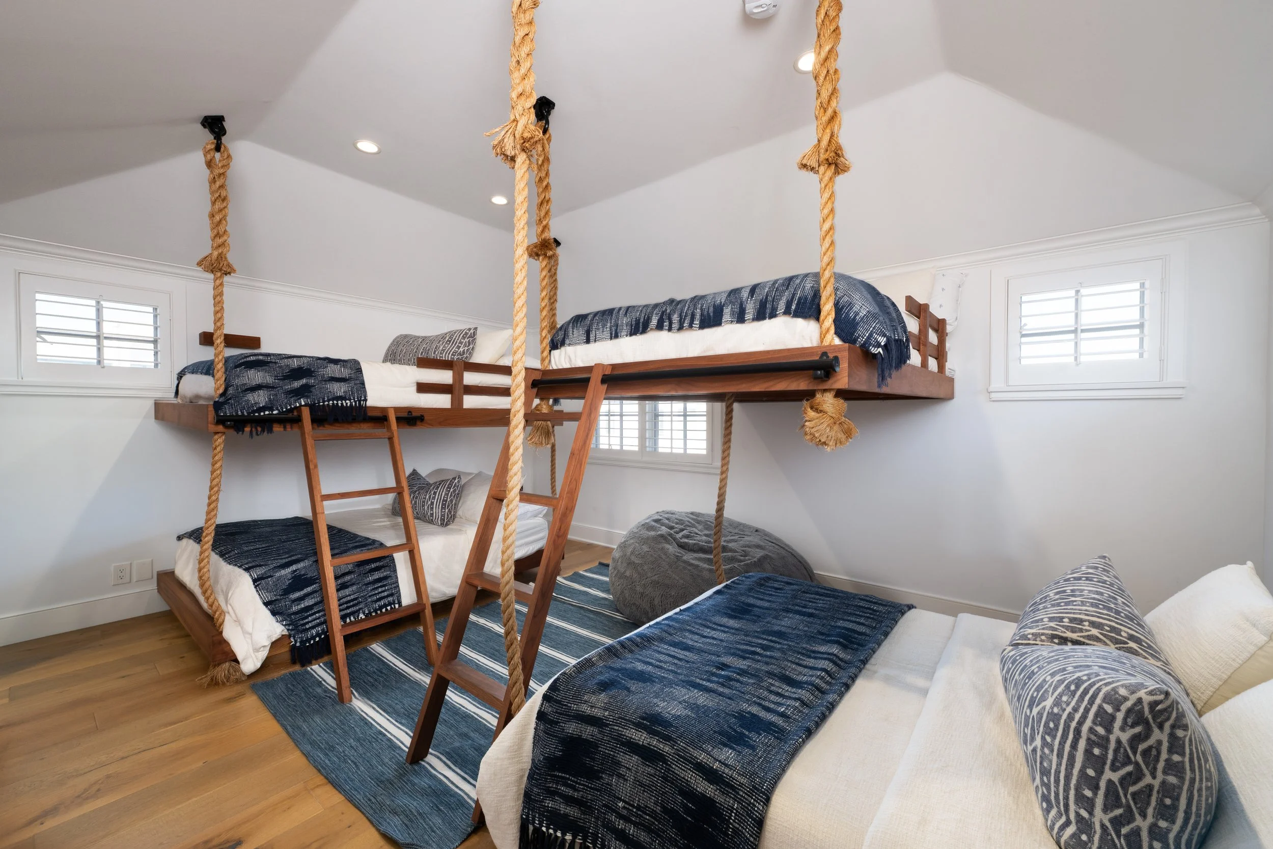 A bedroom with a loft bed made of wood, accessible by a small ladder, with a platform that has a bed with blue and white bedding. Rope supports hold up the loft, and there are windows with shutters on the walls. The room has hardwood floors and a str