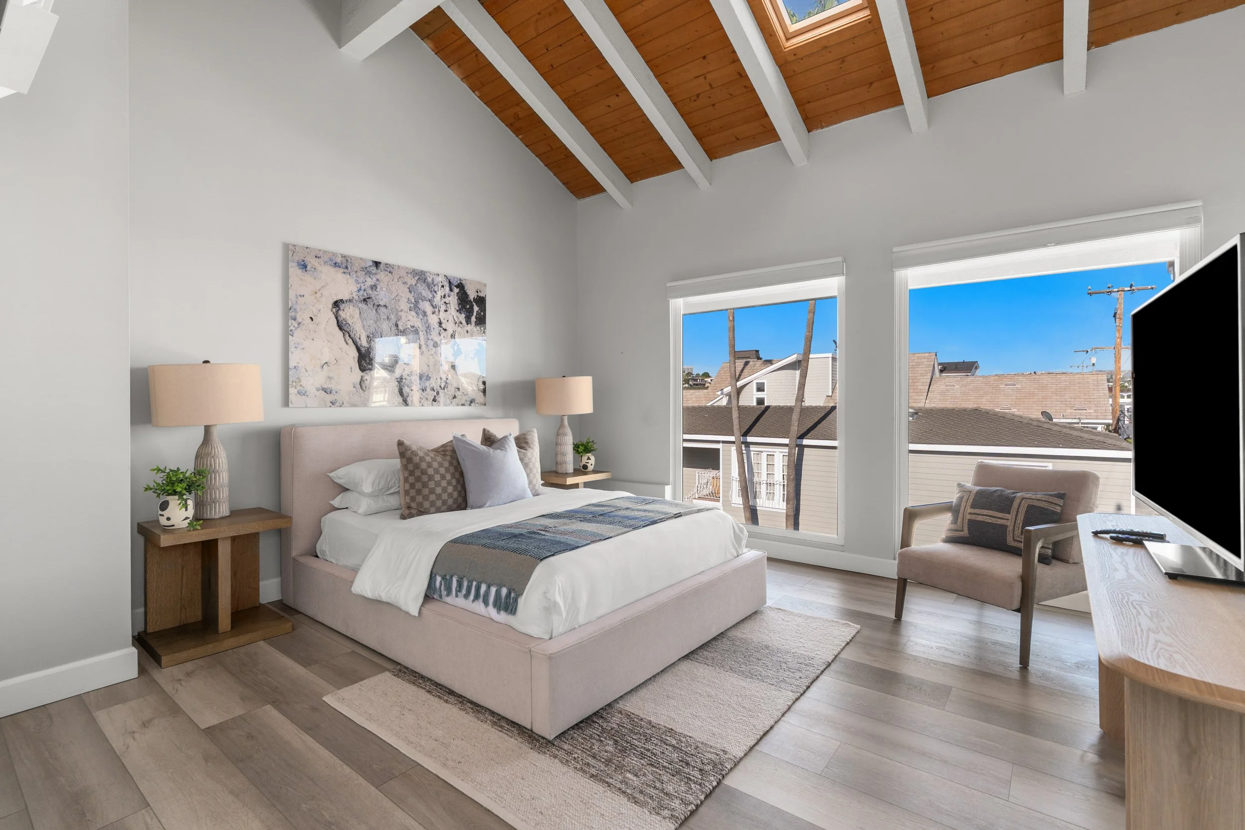 A bedroom with a beige bed, two wooden nightstands with lamps, a painting above the bed, large windows showing a view of rooftops and trees, a beige armchair, and a flat-screen TV on a white dresser. The room has white walls, a wood-paneled ceiling w