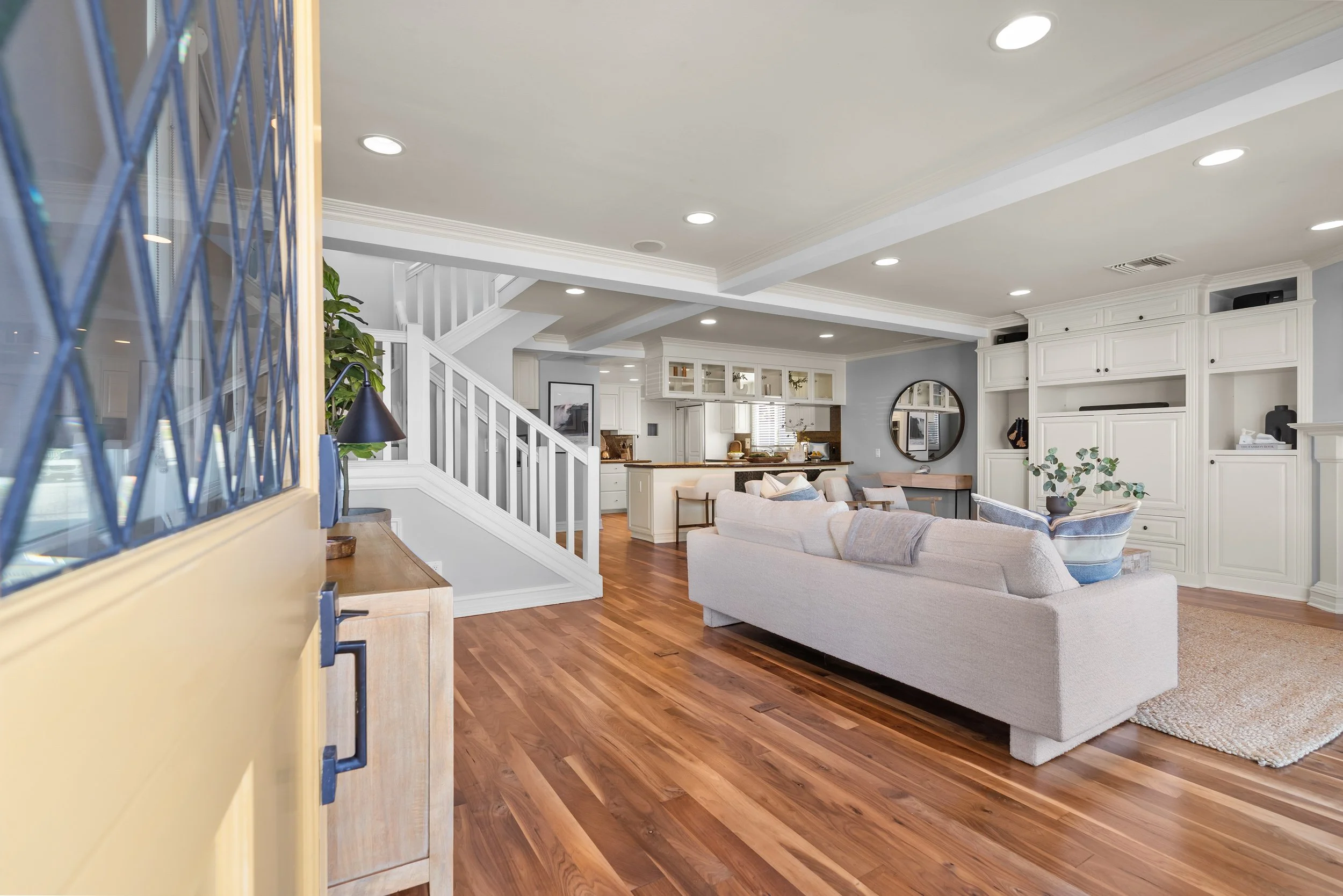 A spacious, modern living room with hardwood floors, white walls, and ceiling with recessed lighting. There are white built-in cabinets, a round mirror, and a cozy seating area with a beige sofa and chairs. A staircase with white railing leads to the