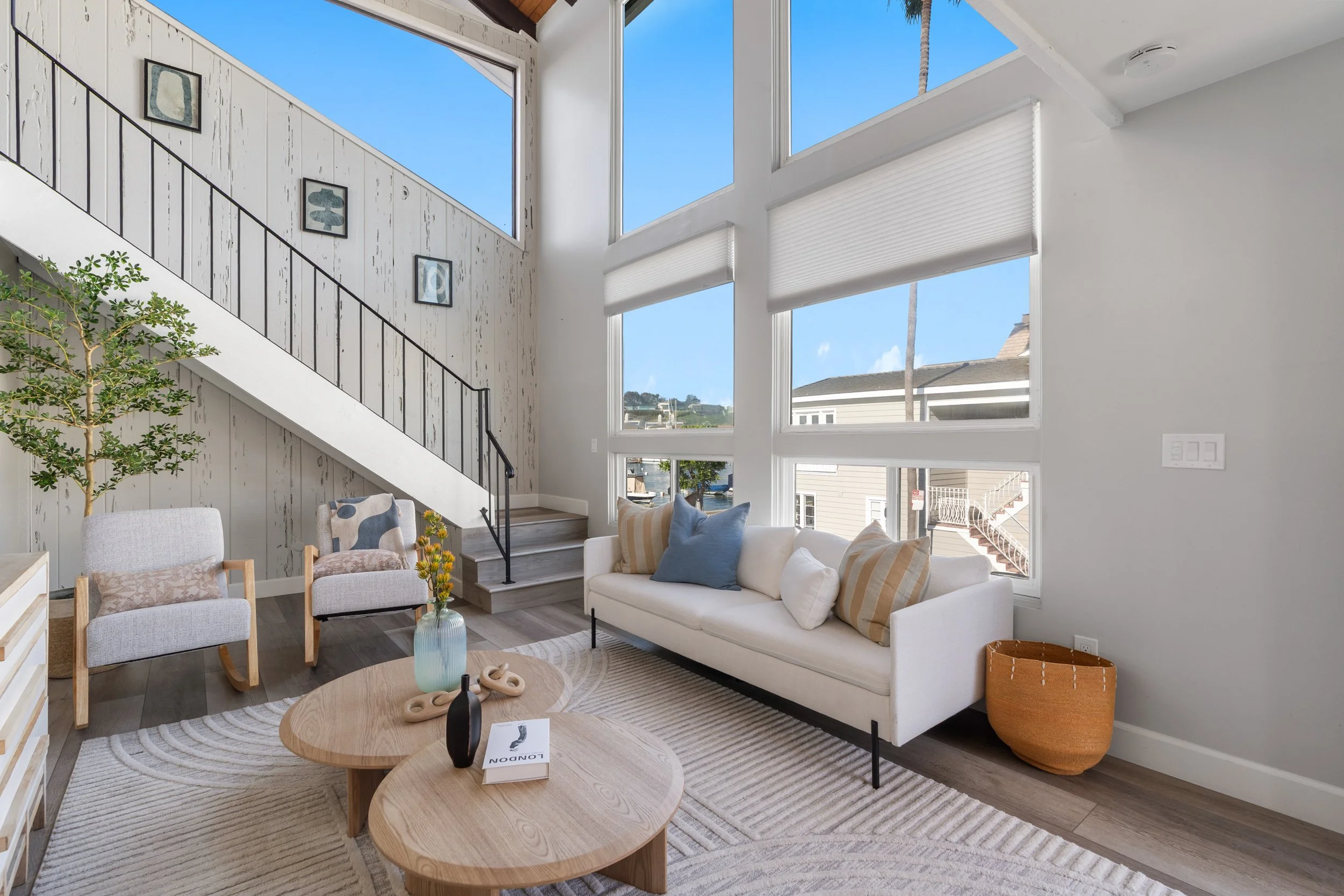 Living room with large windows, white sofa with striped pillows, two beige armchairs, a round wooden coffee table with decorations, a plant, and wall art, with a view of other houses outside.