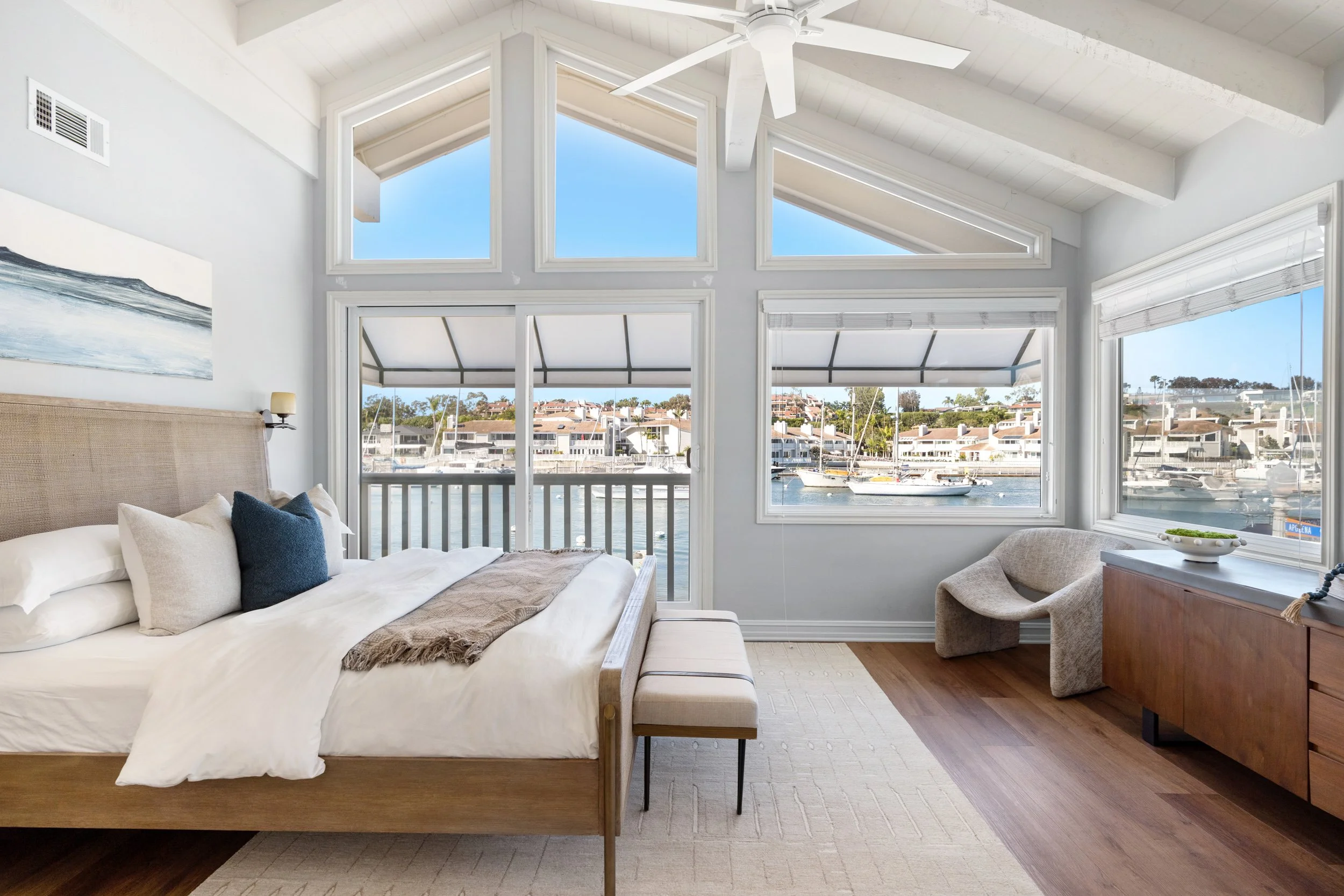 A bedroom with large windows and a view of boats and water outside, a bed with white and navy pillows, a wooden dresser, a beige armchair, and a ceiling fan.