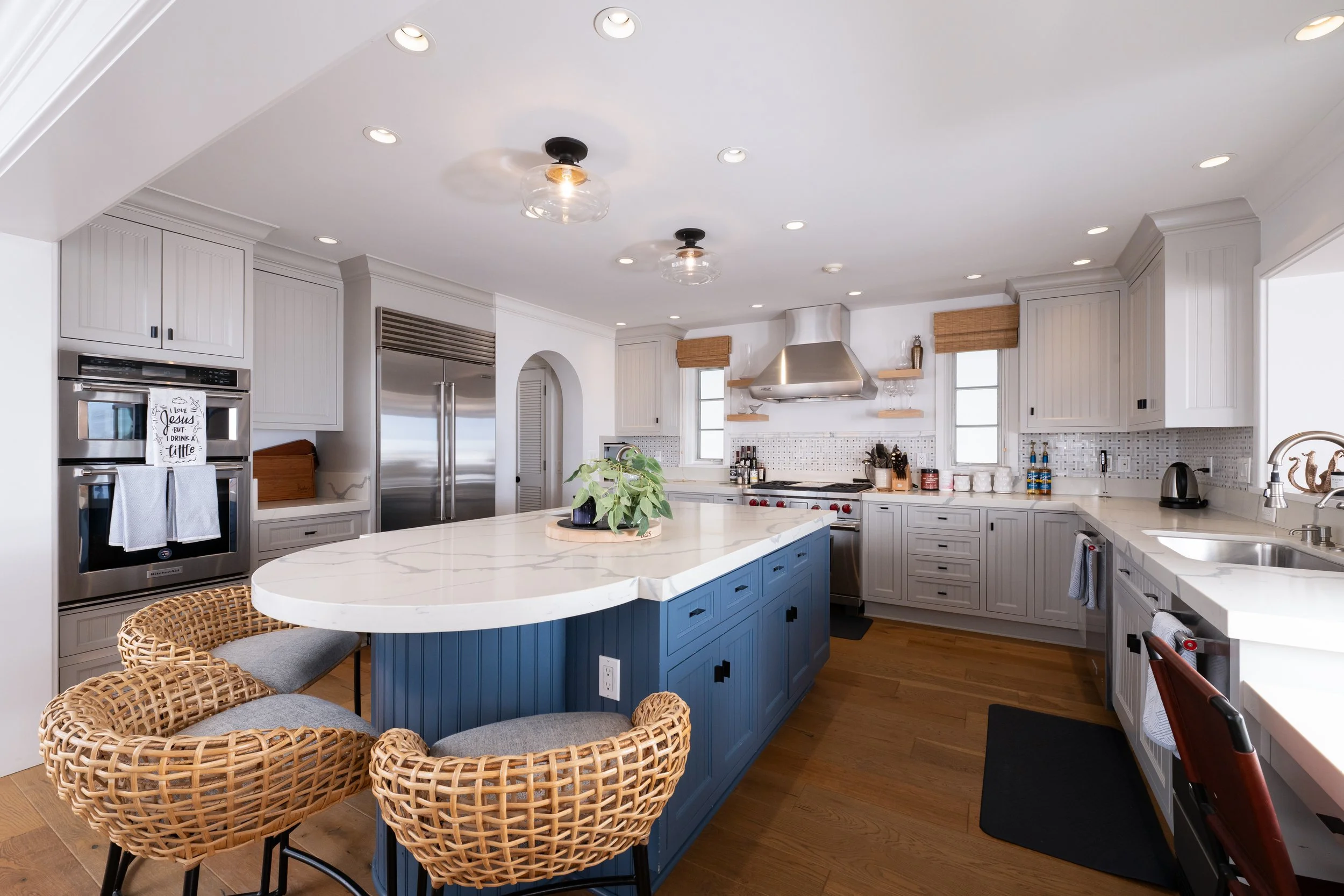 Modern kitchen with white cabinets, stainless steel appliances, a blue island, and woven chairs around a white marble countertop. Recessed lighting and small windows with bamboo shades are also visible.