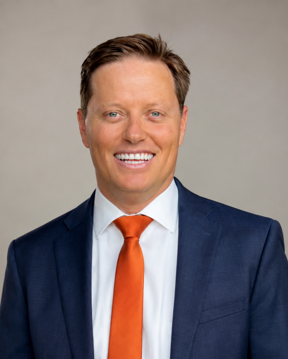 A smiling man wearing a dark blue suit, white shirt, and orange tie against a neutral background.