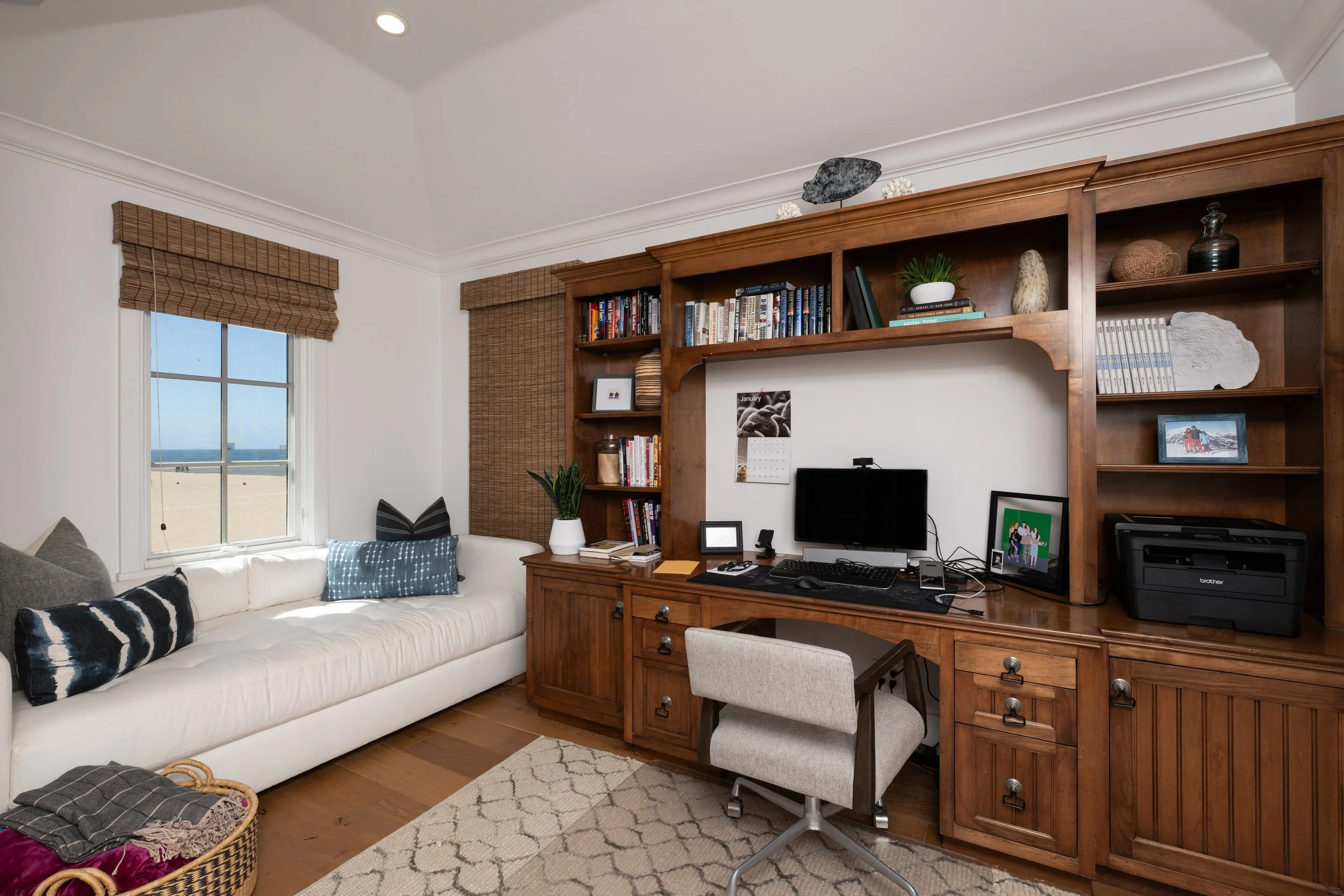 Living room with white sofa with black and white pillows, a large wooden bookshelf with decorative items and books, a desk with a computer, a printer, and framed photos, a window with brown blinds, and a view of the beach outside.