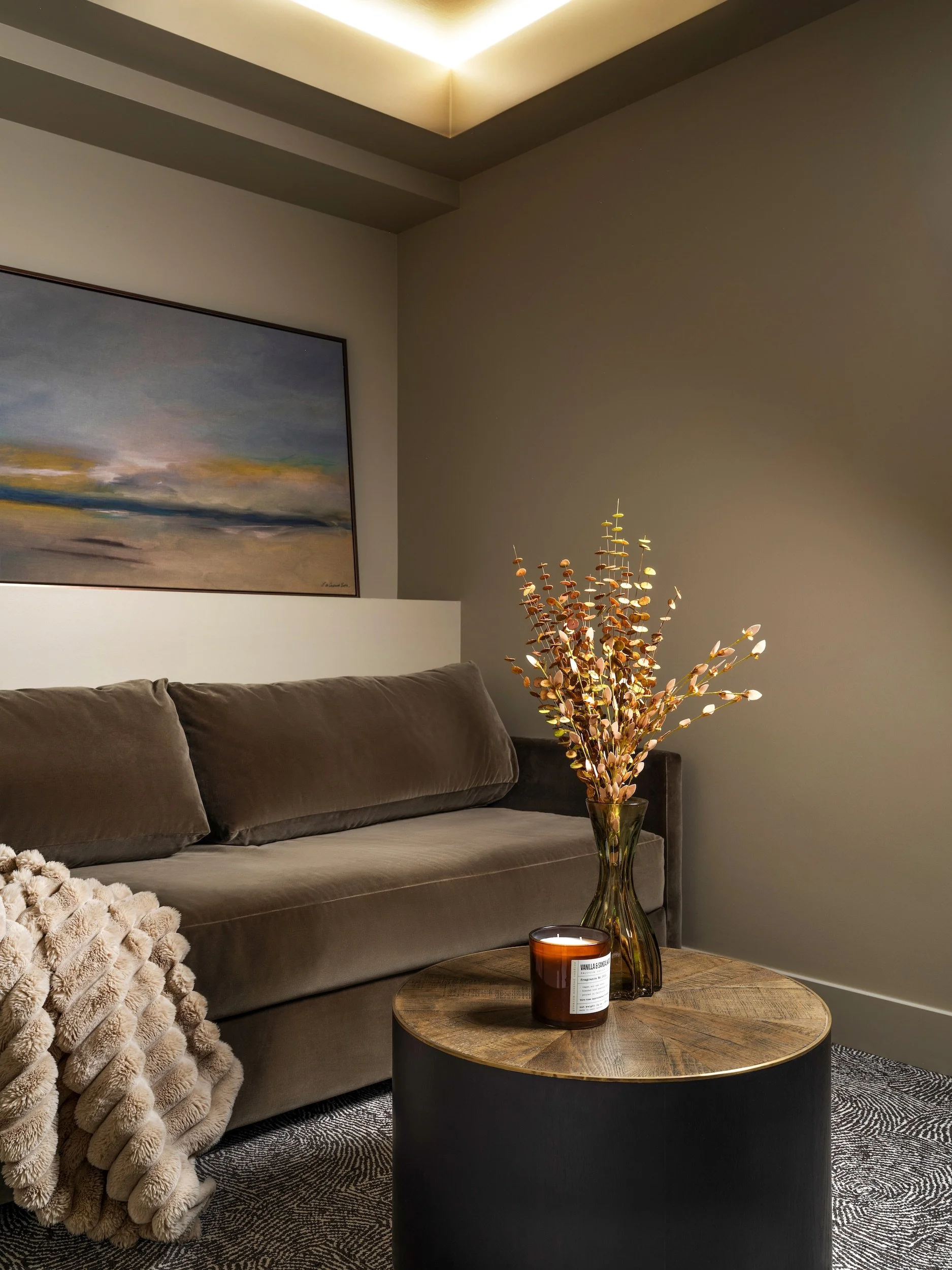 Cozy sitting room corner with taupe velvet sectional, faux fur throw, cylindrical wood and brass side table, dried branch arrangement, and large-scale landscape painting, Atlanta, GA private residence, interior design by Luxe Style Studio
